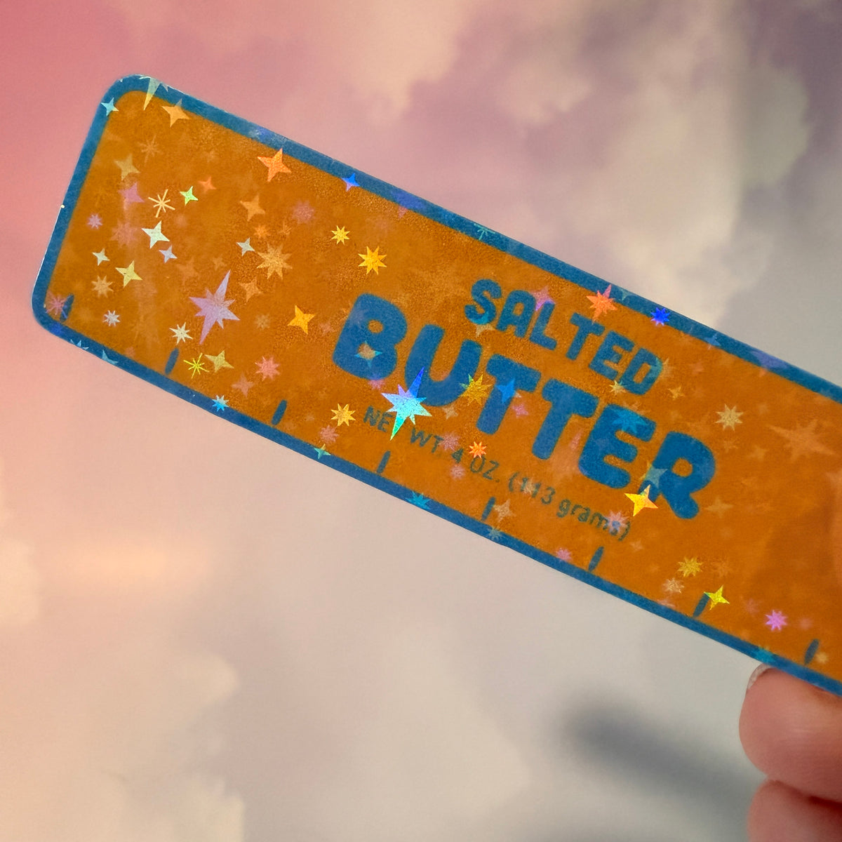 Holographic vinyl sticker shaped like a classic stick of butter, measuring approximately 3 inches long by 1 inch tall. Colorful reflective finish adds a playful touch to laptops, notebooks, tumblers, and sticker collections.