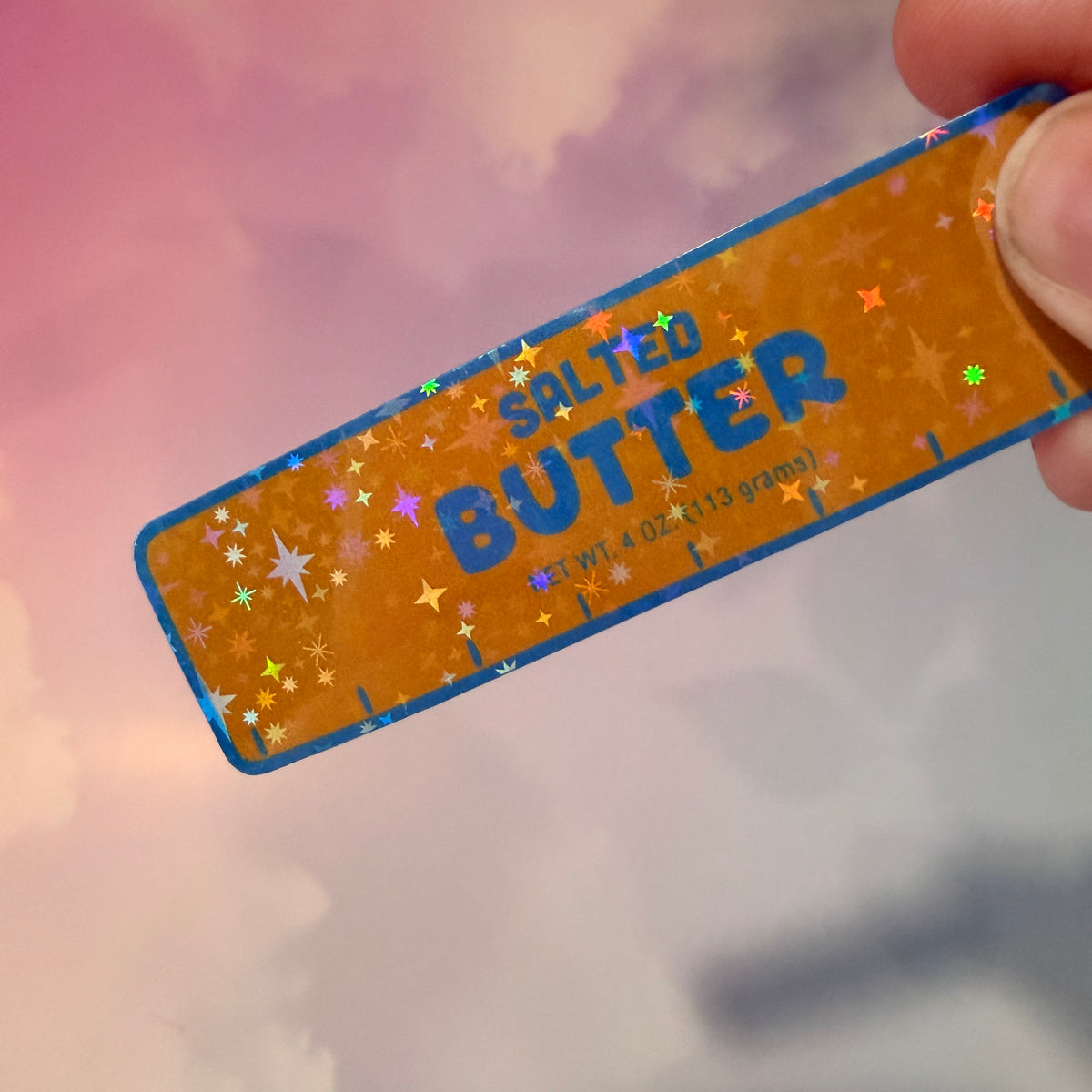 Holographic vinyl sticker shaped like a classic stick of butter, measuring approximately 3 inches long by 1 inch tall. Colorful reflective finish adds a playful touch to laptops, notebooks, tumblers, and sticker collections.