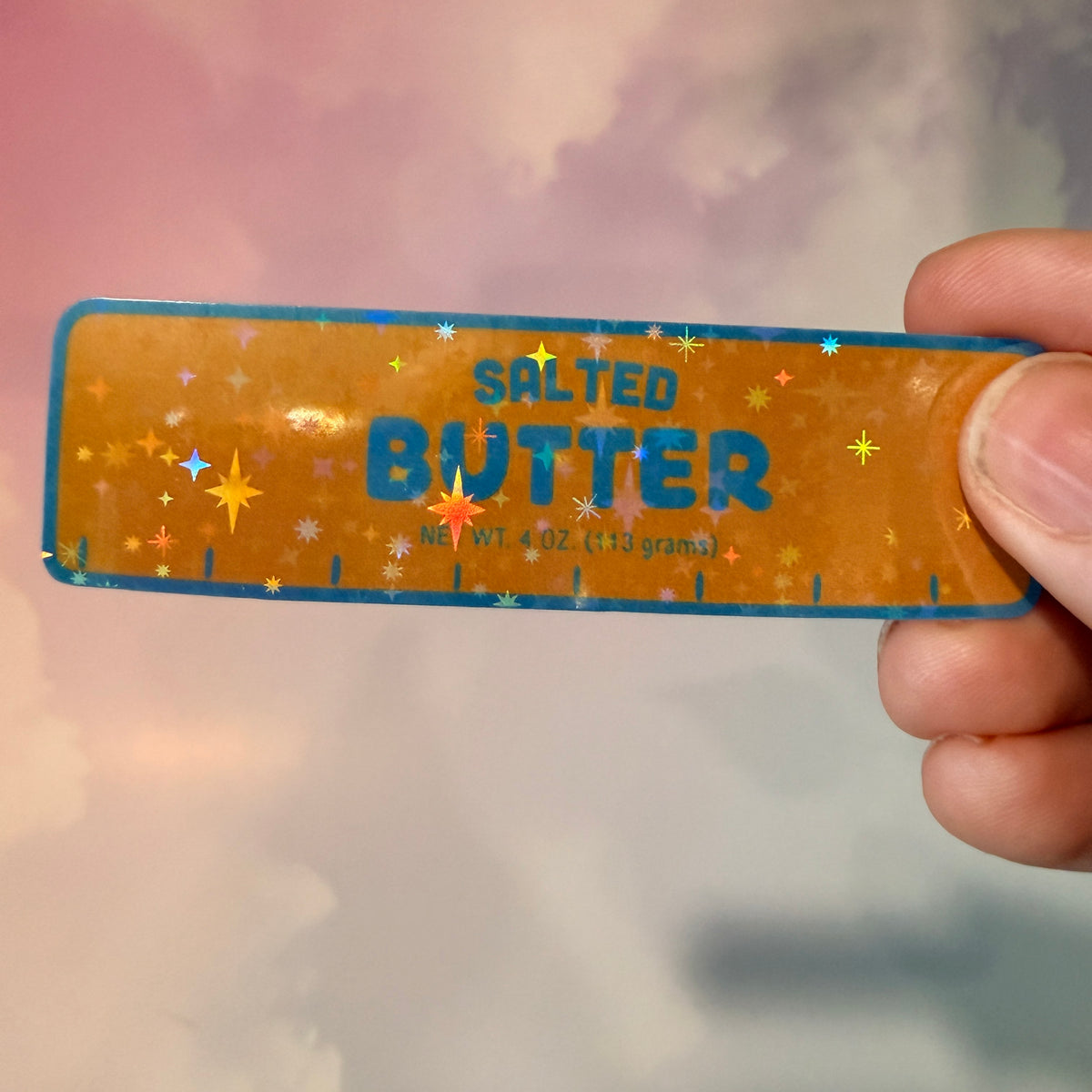 Holographic vinyl sticker shaped like a classic stick of butter, measuring approximately 3 inches long by 1 inch tall. Colorful reflective finish adds a playful touch to laptops, notebooks, tumblers, and sticker collections.