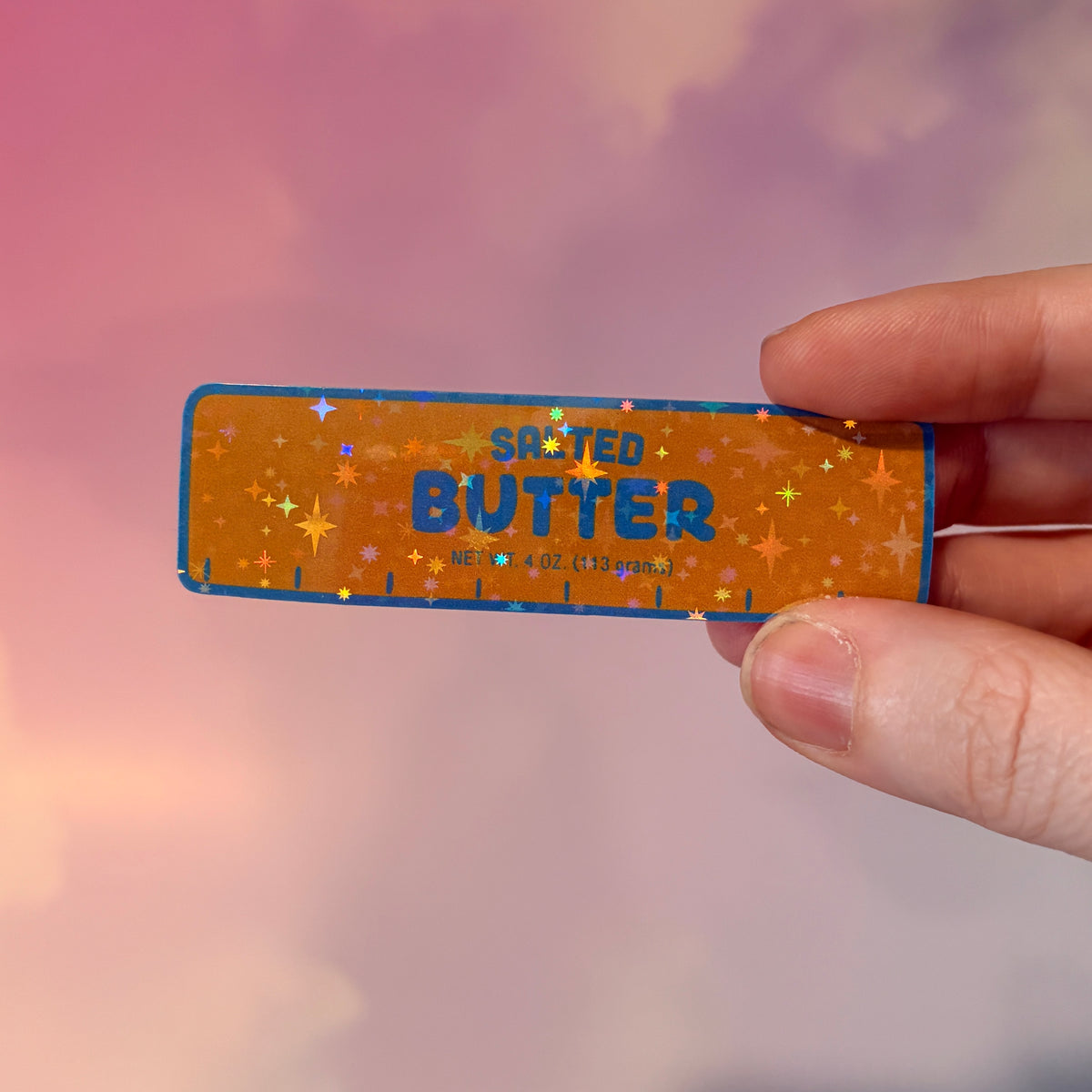 Holographic vinyl sticker shaped like a classic stick of butter, measuring approximately 3 inches long by 1 inch tall. Colorful reflective finish adds a playful touch to laptops, notebooks, tumblers, and sticker collections.