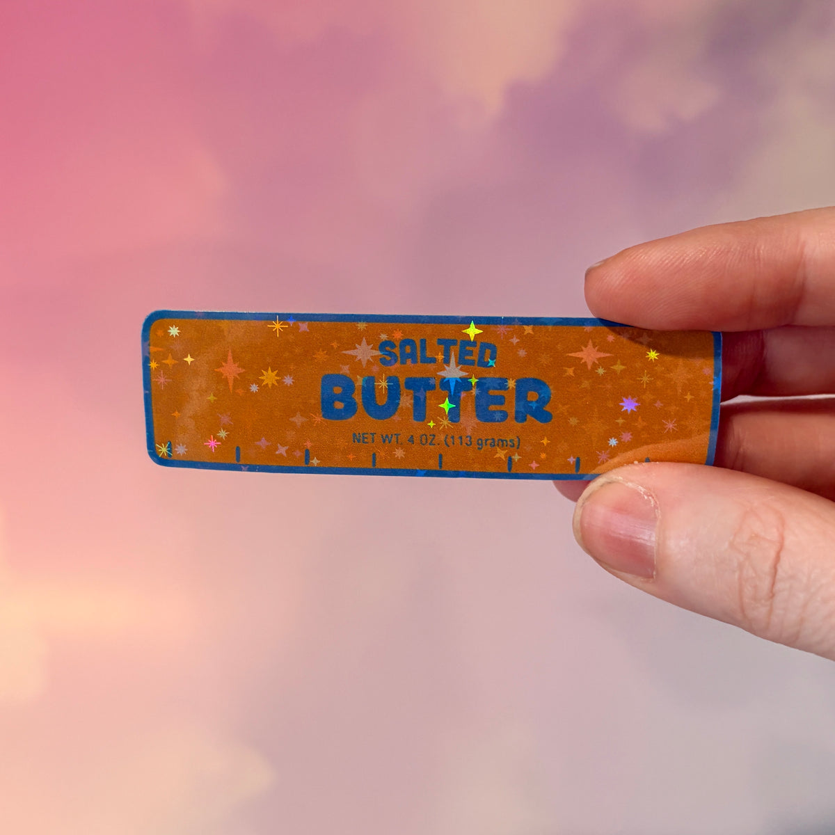 Holographic vinyl sticker shaped like a classic stick of butter, measuring approximately 3 inches long by 1 inch tall. Colorful reflective finish adds a playful touch to laptops, notebooks, tumblers, and sticker collections.