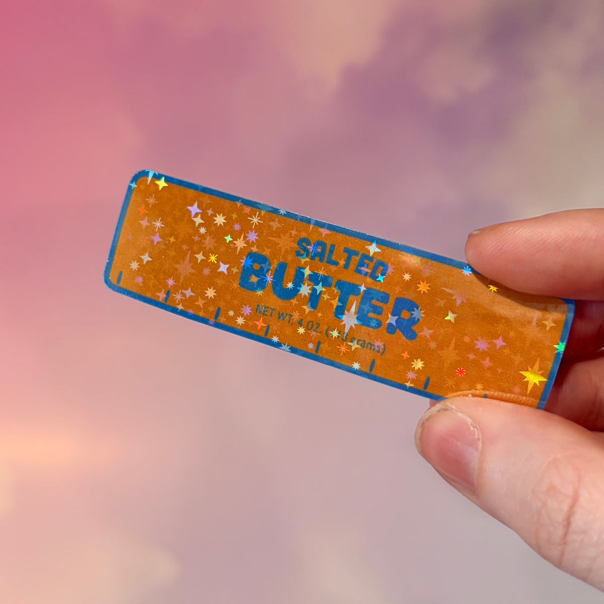 Holographic vinyl sticker shaped like a classic stick of butter, measuring approximately 3 inches long by 1 inch tall. Colorful reflective finish adds a playful touch to laptops, notebooks, tumblers, and sticker collections.