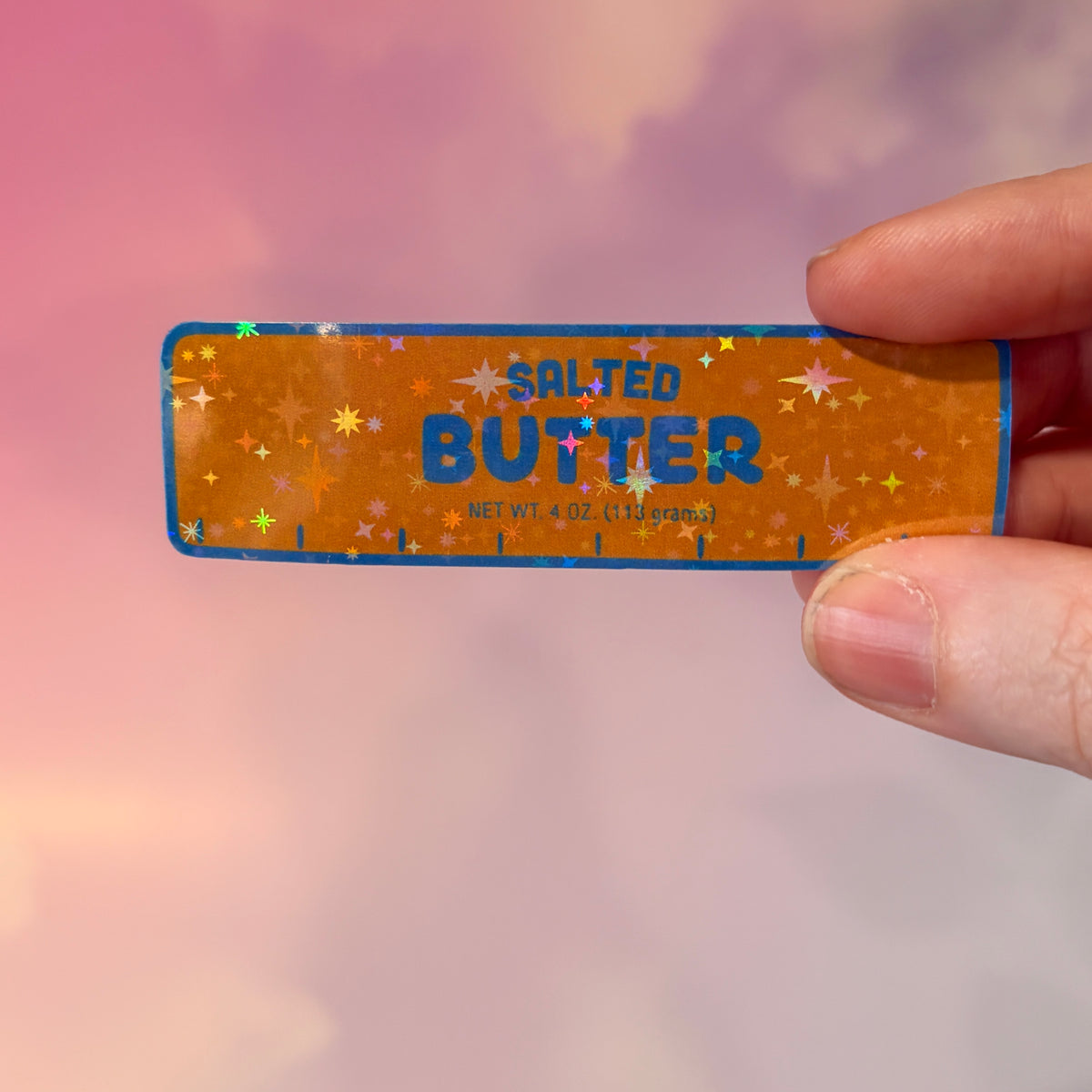 Holographic vinyl sticker shaped like a classic stick of butter, measuring approximately 3 inches long by 1 inch tall. Colorful reflective finish adds a playful touch to laptops, notebooks, tumblers, and sticker collections.