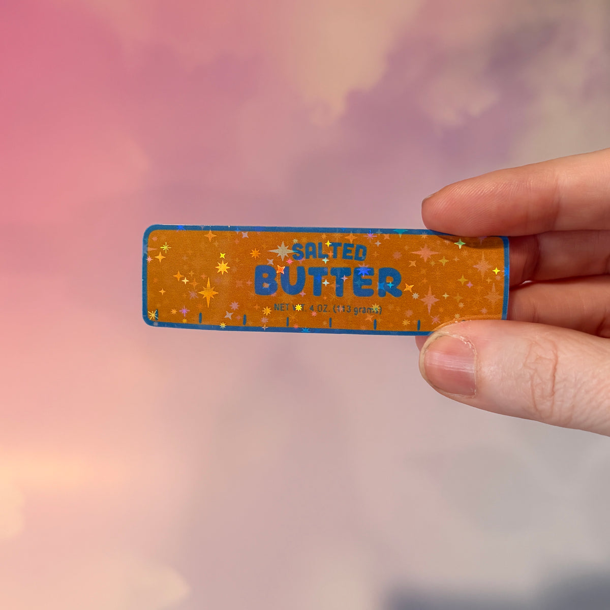 Holographic vinyl sticker shaped like a classic stick of butter, measuring approximately 3 inches long by 1 inch tall. Colorful reflective finish adds a playful touch to laptops, notebooks, tumblers, and sticker collections.