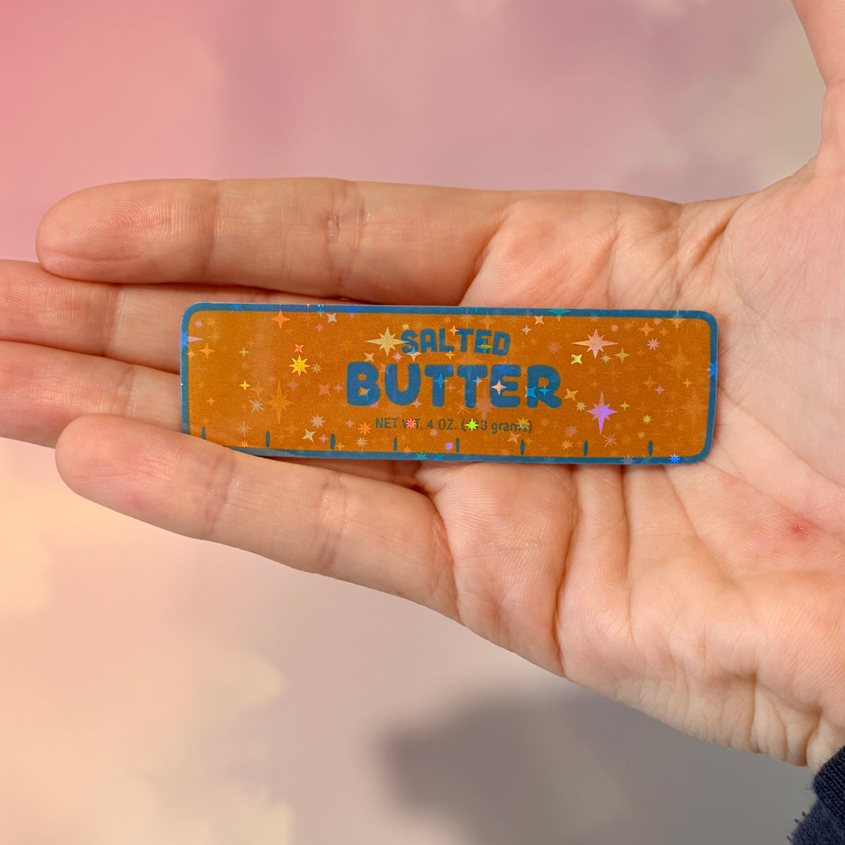 Holographic vinyl sticker shaped like a classic stick of butter, measuring approximately 3 inches long by 1 inch tall. Colorful reflective finish adds a playful touch to laptops, notebooks, tumblers, and sticker collections.