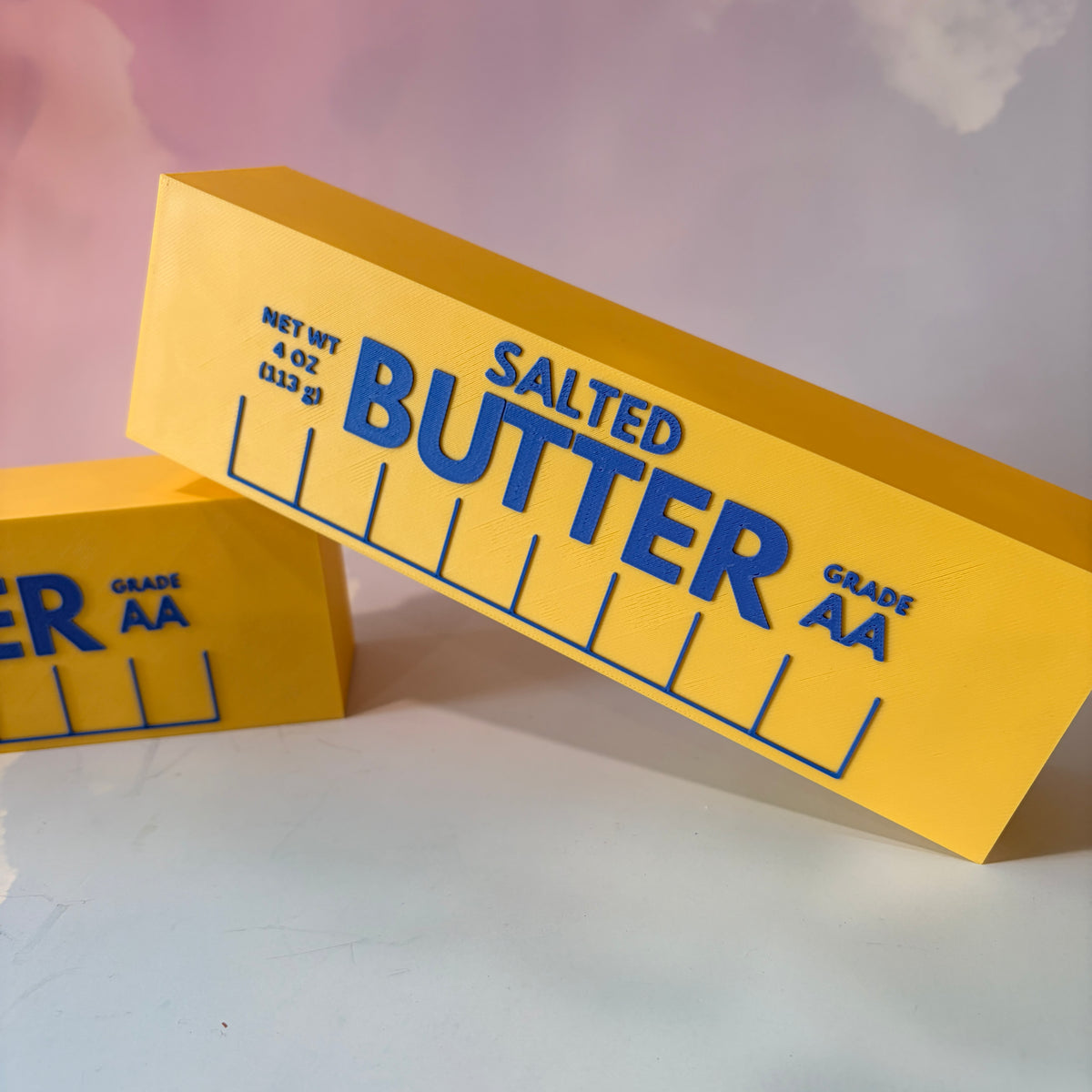 Oversized 3D printed novelty stick of butter, approximately 8.5 inches long, featuring classic tablespoon measurement lines, Grade AA labeling, and net weight details. Whimsical food-themed decor for kitchens and farmhouse-style homes.
