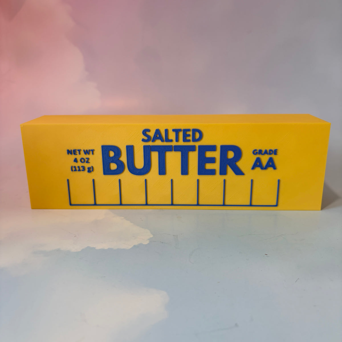 Oversized 3D printed novelty stick of butter, approximately 8.5 inches long, featuring classic tablespoon measurement lines, Grade AA labeling, and net weight details. Whimsical food-themed decor for kitchens and farmhouse-style homes.