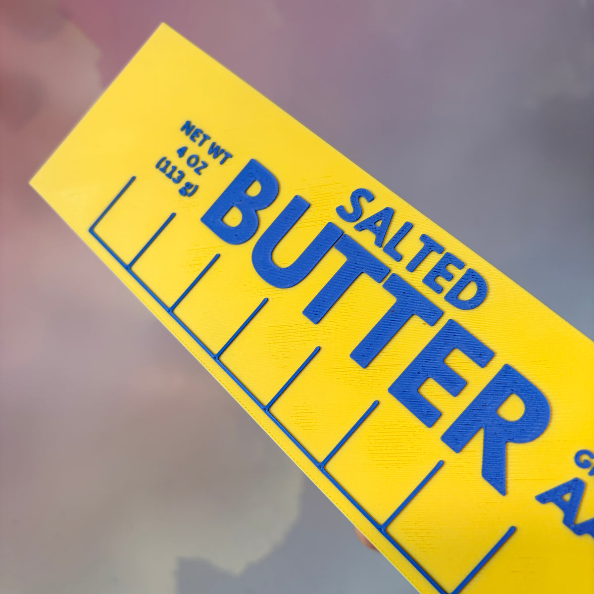 Oversized 3D printed novelty stick of butter, approximately 8.5 inches long, featuring classic tablespoon measurement lines, Grade AA labeling, and net weight details. Whimsical food-themed decor for kitchens and farmhouse-style homes.