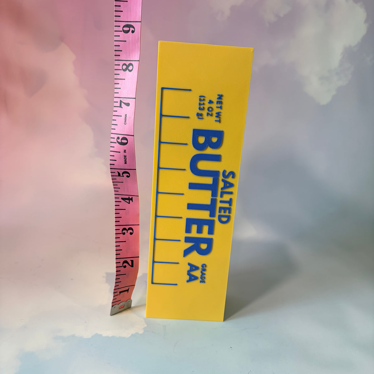 Oversized 3D printed novelty stick of butter, approximately 8.5 inches long, featuring classic tablespoon measurement lines, Grade AA labeling, and net weight details. Whimsical food-themed decor for kitchens and farmhouse-style homes.