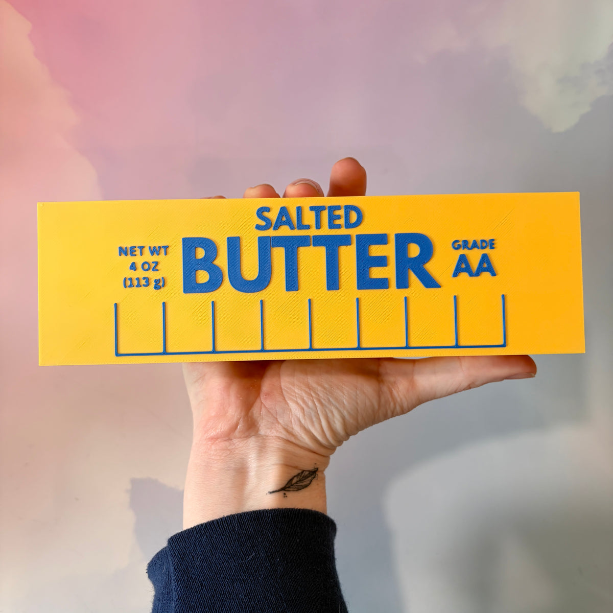 Oversized 3D printed novelty stick of butter, approximately 8.5 inches long, featuring classic tablespoon measurement lines, Grade AA labeling, and net weight details. Whimsical food-themed decor for kitchens and farmhouse-style homes.