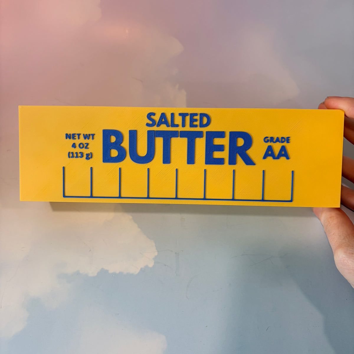 Oversized 3D printed novelty stick of butter, approximately 8.5 inches long, featuring classic tablespoon measurement lines, Grade AA labeling, and net weight details. Whimsical food-themed decor for kitchens and farmhouse-style homes.