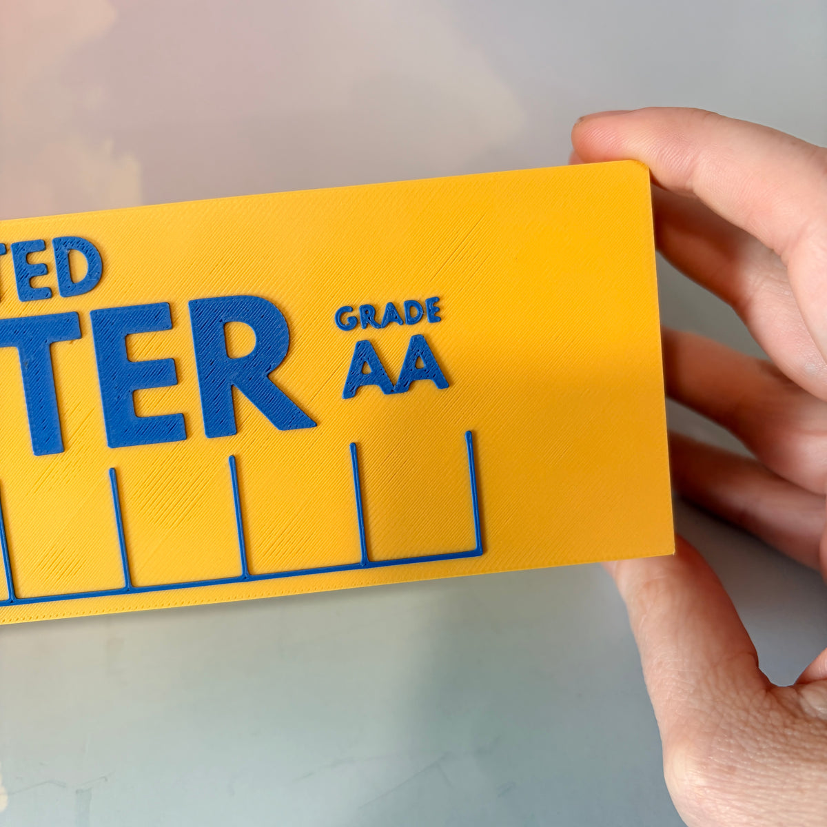 Oversized 3D printed novelty stick of butter, approximately 8.5 inches long, featuring classic tablespoon measurement lines, Grade AA labeling, and net weight details. Whimsical food-themed decor for kitchens and farmhouse-style homes.