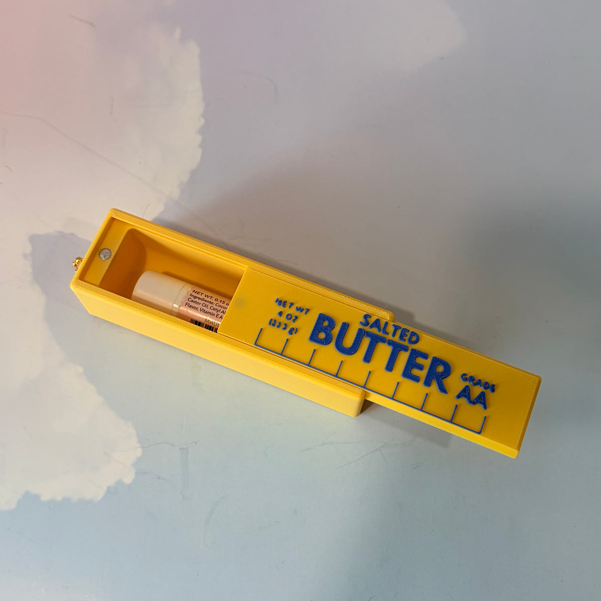 3D printed stick of butter bag charm with gold hardware, classic TBSP markings, Grade AA label, and net weight details. Sliding top hides a lipstick or chapstick in a magnetic compartment.