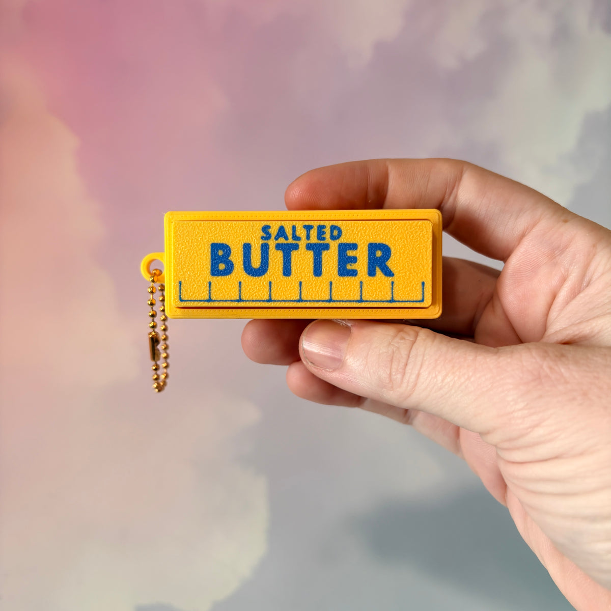 3D printed stick of butter fidget clicker keychain with classic tablespoon marking lines, gold-tone hardware, and two satisfying clicky key switches, perfect for foodies and whimsical accessory lovers.