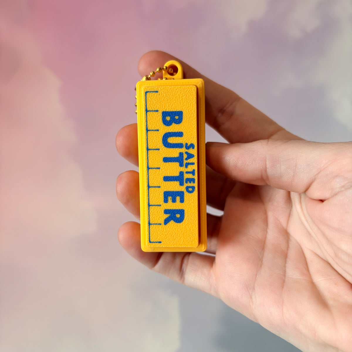 3D printed stick of butter fidget clicker keychain with classic tablespoon marking lines, gold-tone hardware, and two satisfying clicky key switches, perfect for foodies and whimsical accessory lovers.