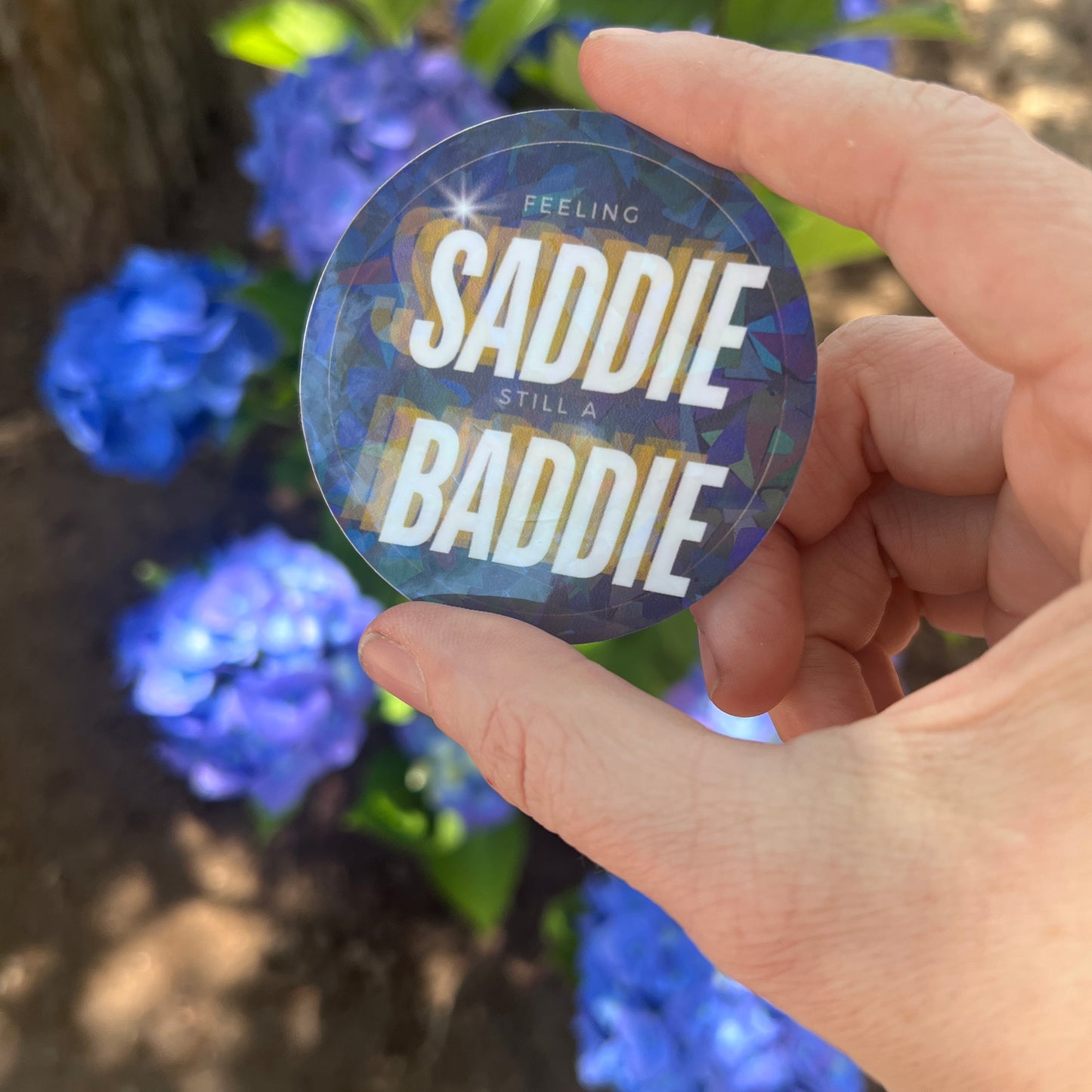 Round holographic sticker with the phrase "Feeling Saddie, Still a Baddie" held in a hand with blue hydrangeas in the background.