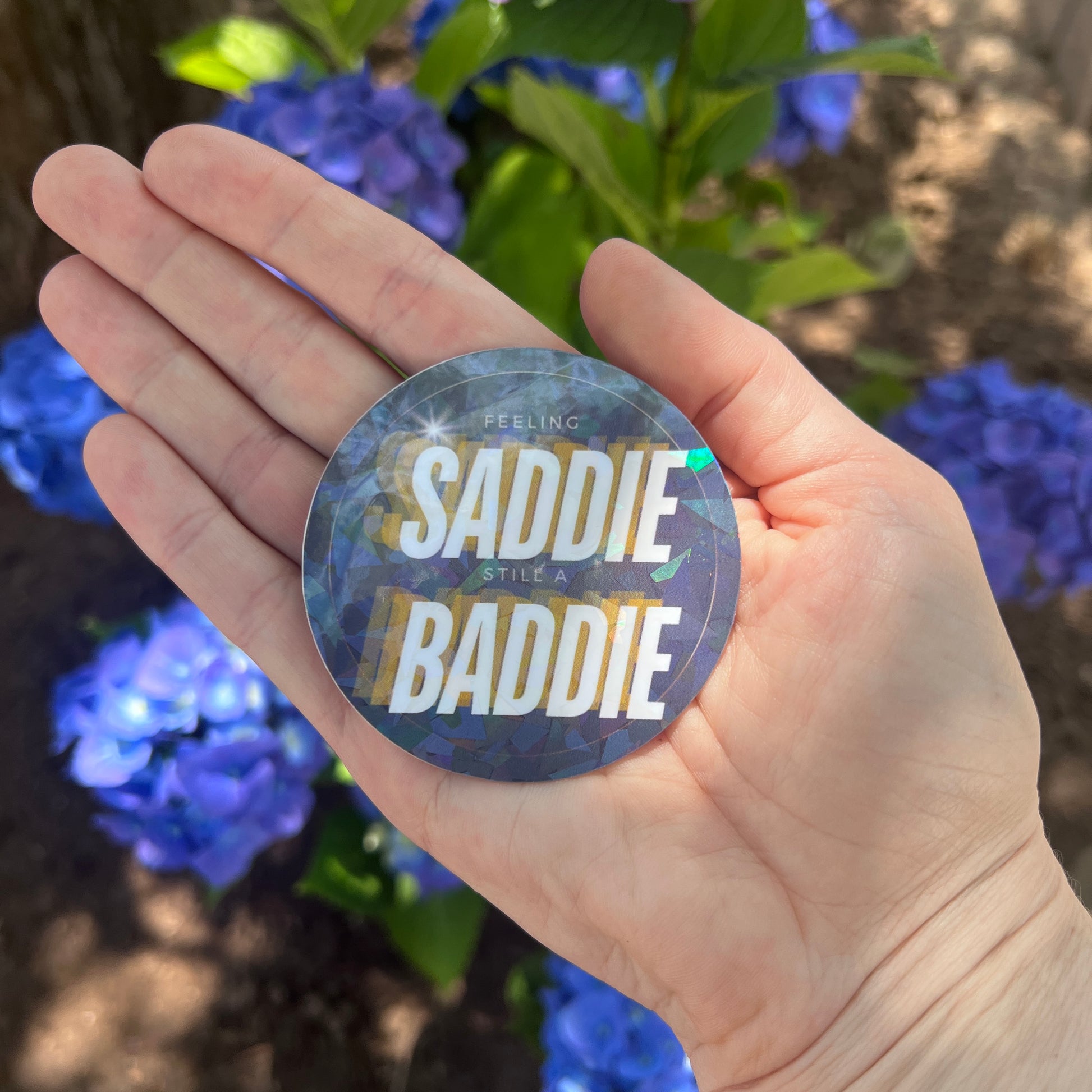 Round holographic sticker with the phrase "Feeling Saddie, Still a Baddie" held in a hand with blue hydrangeas in the background.