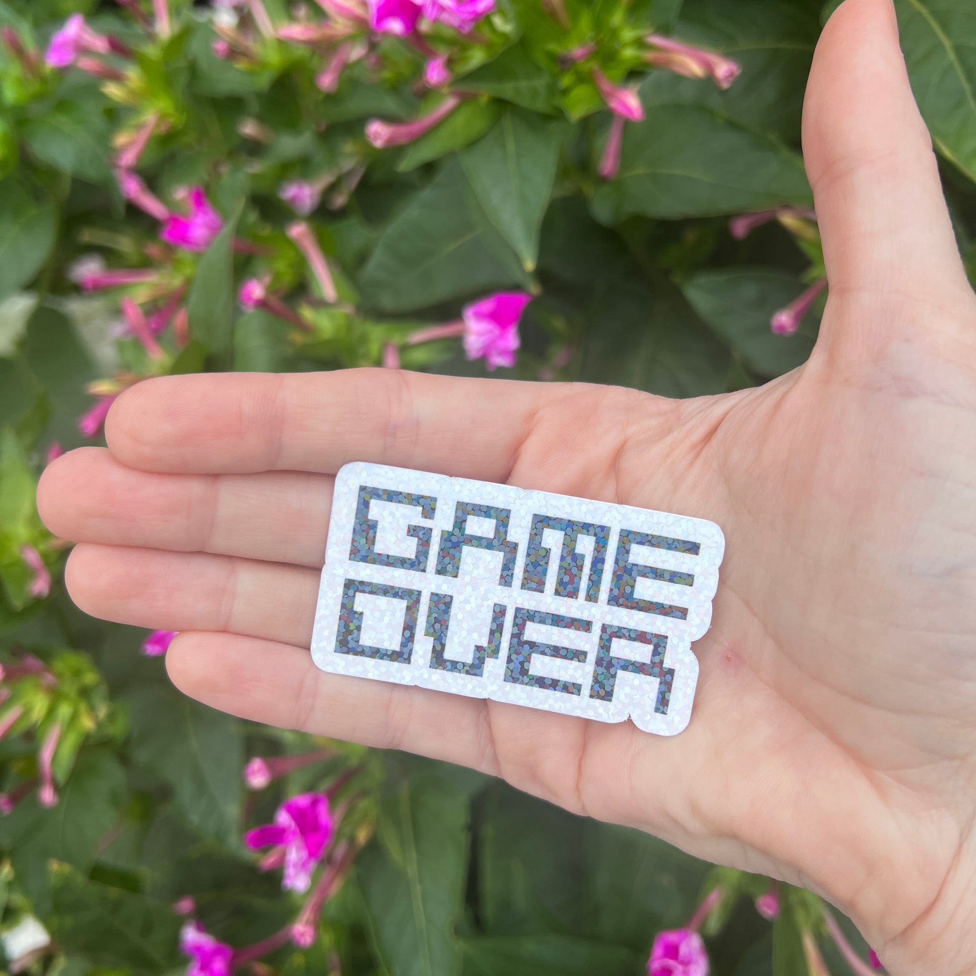 Holographic black and white gamer sticker that says GAME OVER in pixelated retro font. Water-resistant vinyl decal for laptops, bottles, consoles.