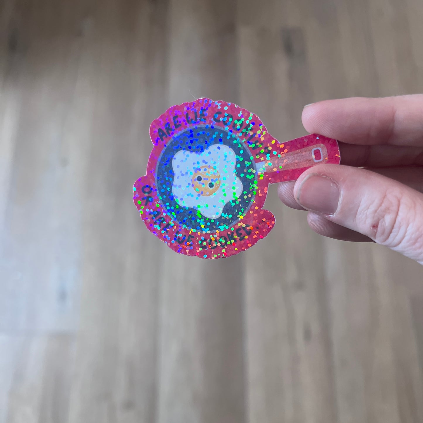 Holographic vinyl sticker with a wide-eyed egg in a frying pan and the phrase “Are We Cooking or Are We Cooked?” Funny kitchen-themed design.