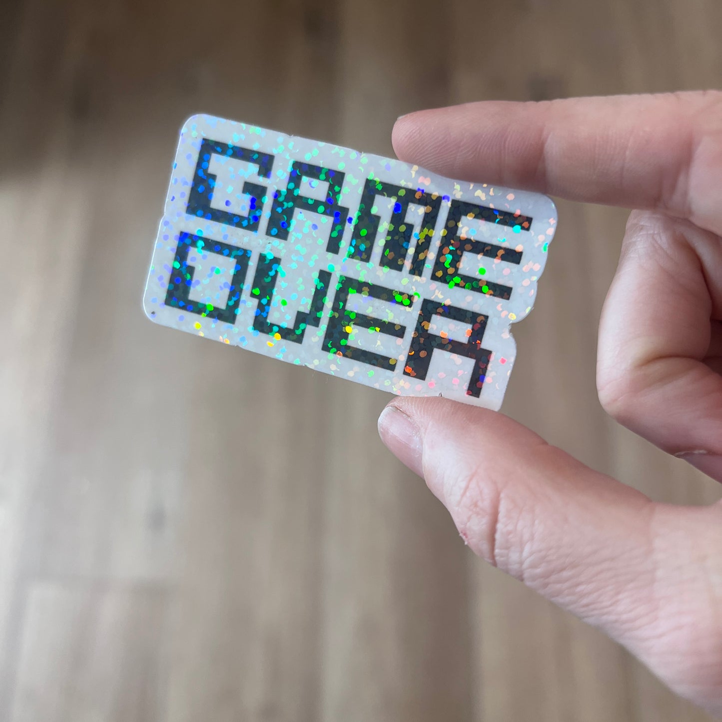 Holographic black and white gamer sticker that says GAME OVER in pixelated retro font. Water-resistant vinyl decal for laptops, bottles, consoles.