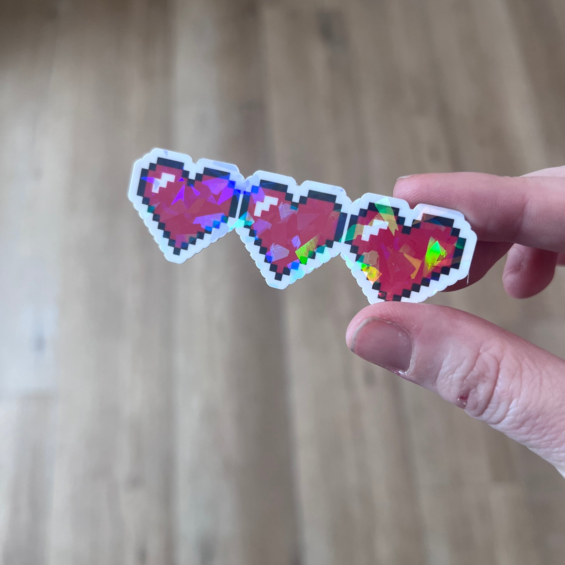 Holographic gamer heart sticker featuring 3 red pixel hearts in a heart container design. Perfect for laptops, water bottles, and game-themed journals.