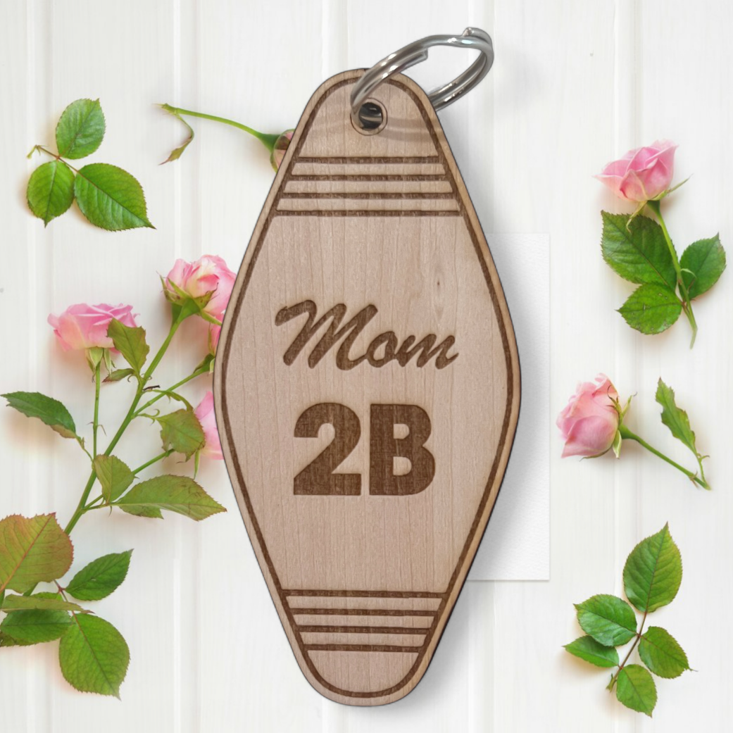 Wooden keychain with 'Mom 2B' text on a white background with floral elements.