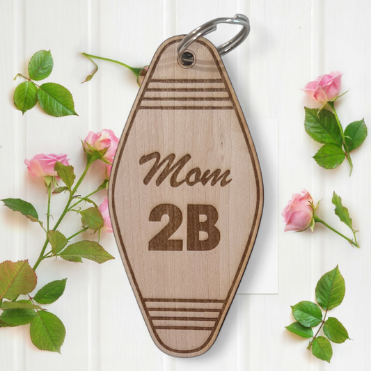 Wooden keychain with 'Mom 2B' text on a white background with floral elements.