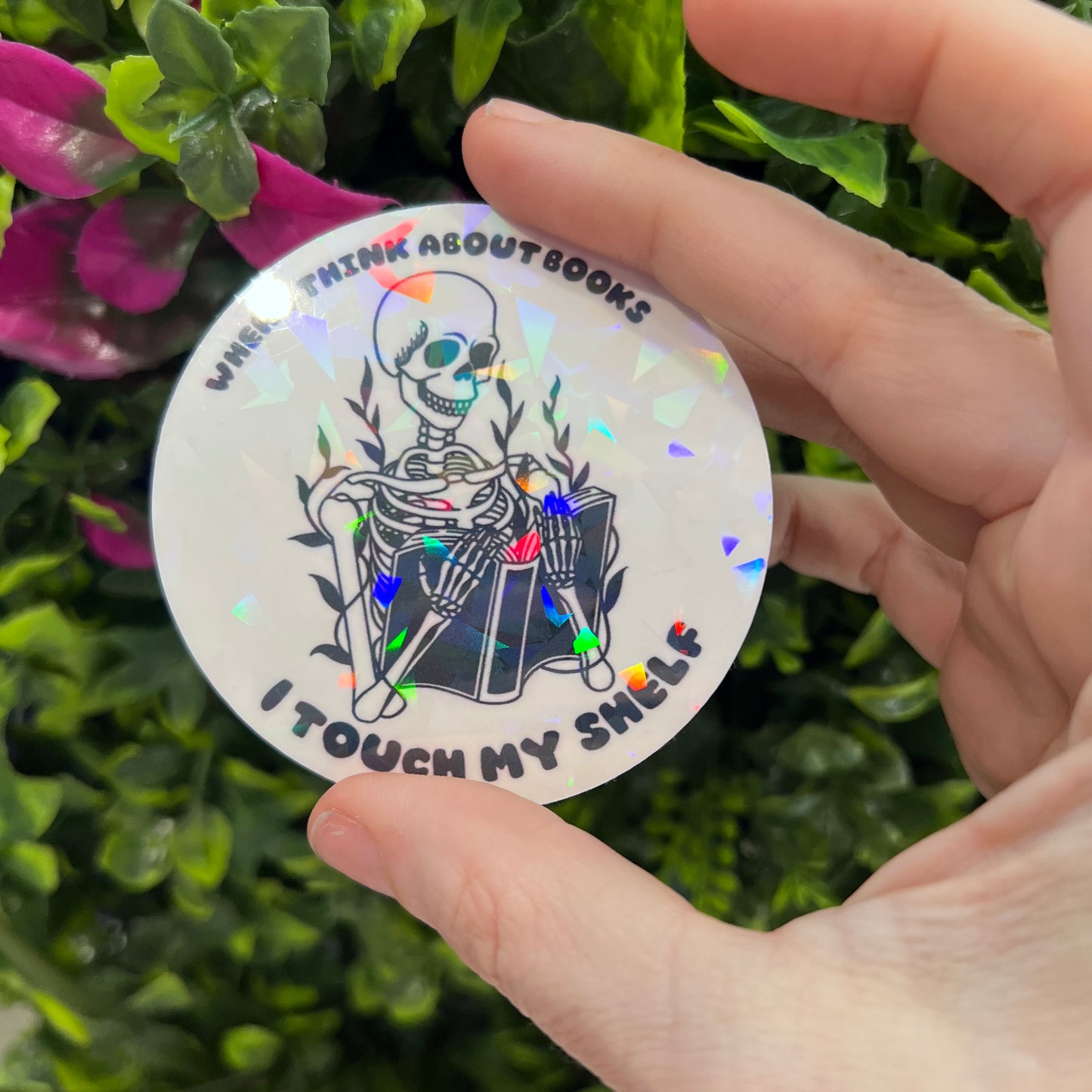 Holographic vinyl sticker with a skeleton reading a book and the phrase "When I think about books I touch my shelf." Perfect for readers and book lovers.