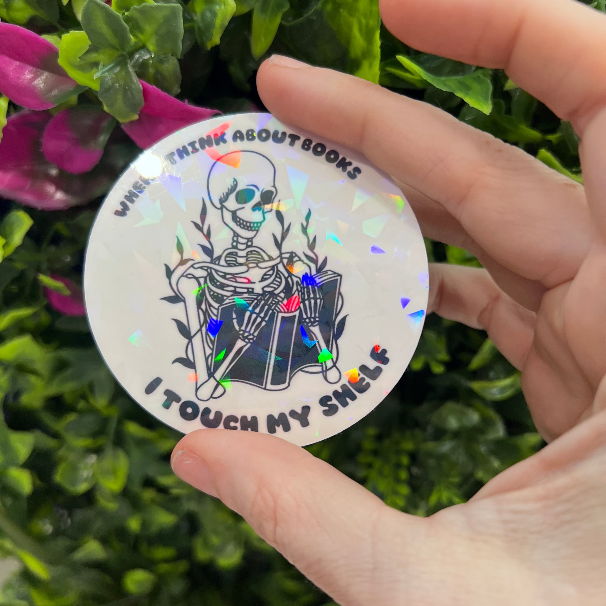 Holographic vinyl sticker with a skeleton reading a book and the phrase "When I think about books I touch my shelf." Perfect for readers and book lovers.