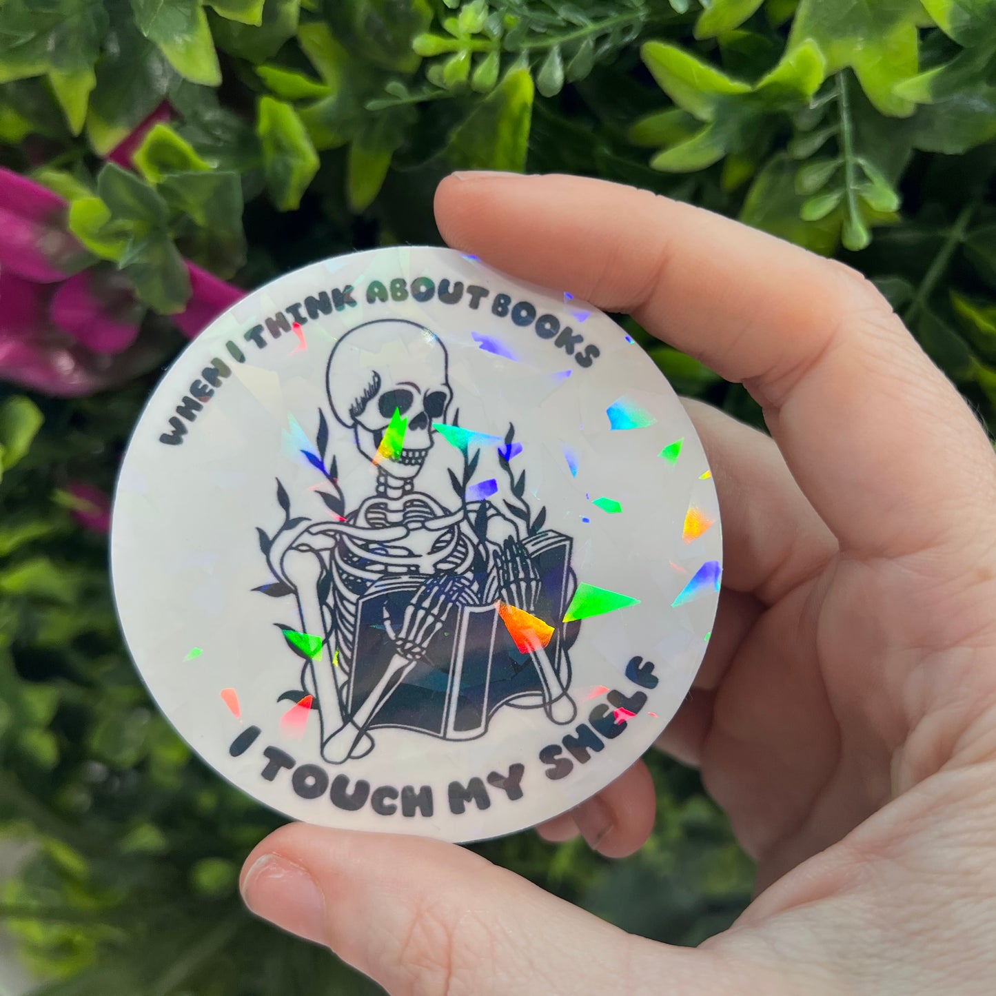 Holographic vinyl sticker with a skeleton reading a book and the phrase "When I think about books I touch my shelf." Perfect for readers and book lovers.