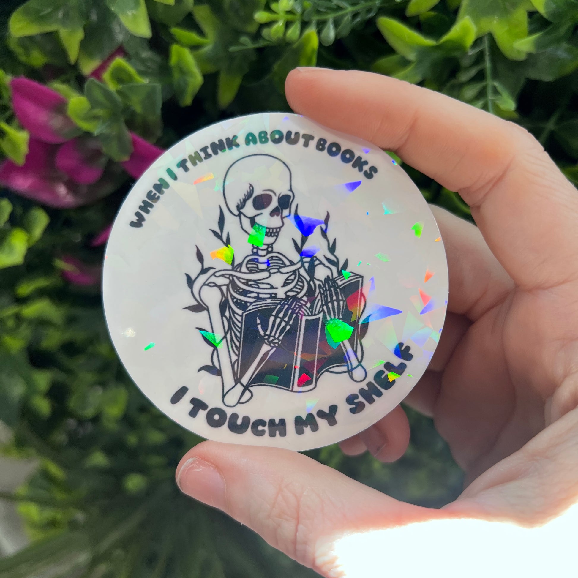 Holographic vinyl sticker with a skeleton reading a book and the phrase "When I think about books I touch my shelf." Perfect for readers and book lovers.