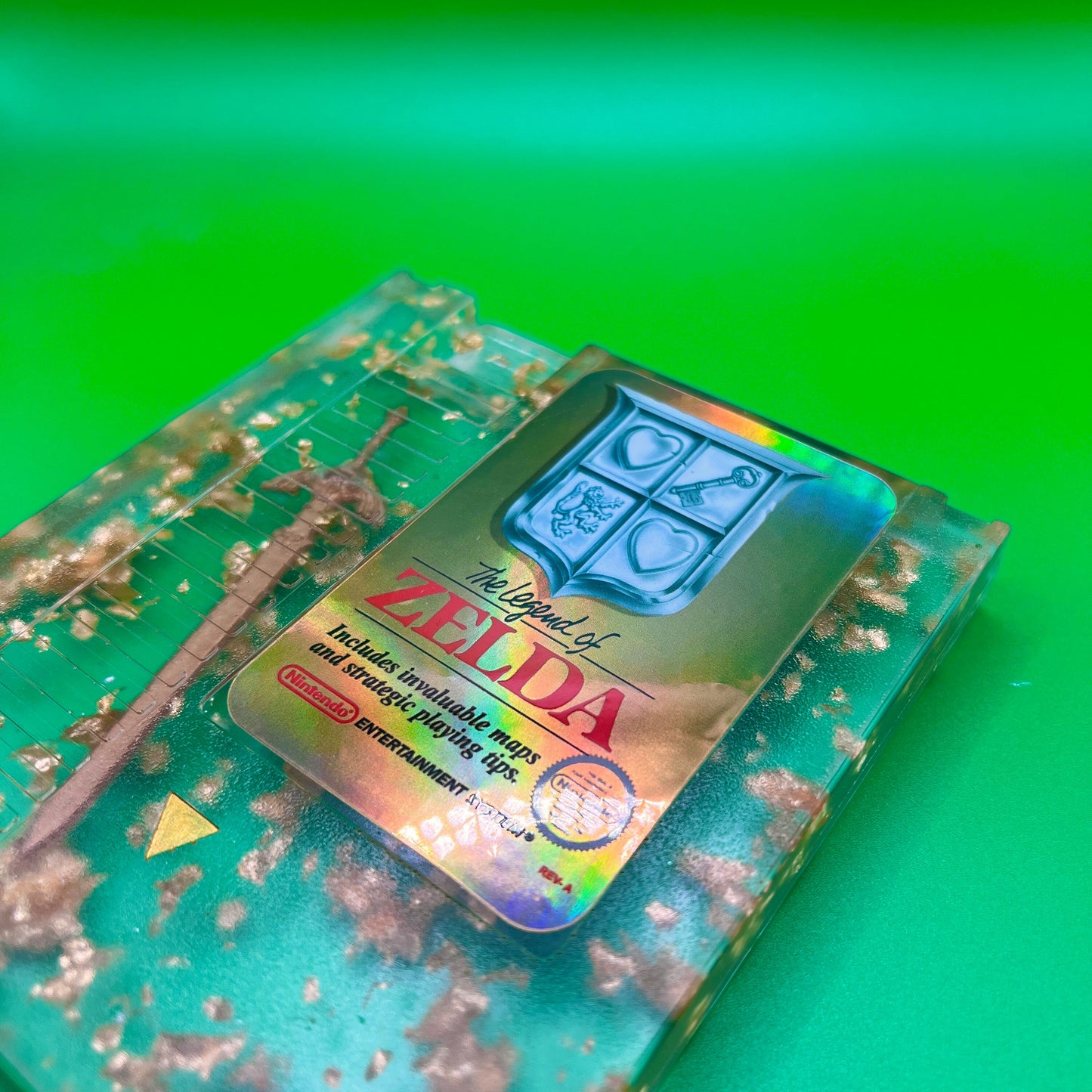 Legend of Zelda resin art featuring a retro NES cartridge with a gold sword design and gold flakes embedded. This handmade display is perfect for gamers and video game collectors, offering a unique décor piece for any gaming setup or retro collection