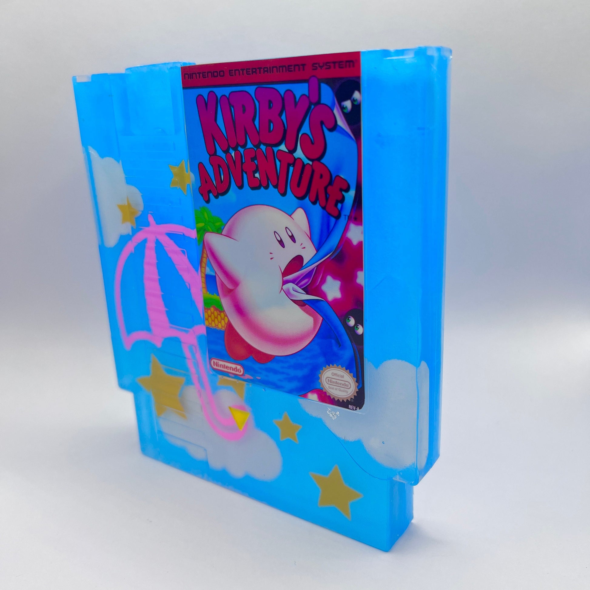 Custom NES cartridge featuring Kirby's Adventure with a colorful design. The blue cartridge has pink umbrellas, clouds, and yellow stars, adding a playful touch for retro gaming fans and collectors. Perfect for showcasing classic Nintendo games.
