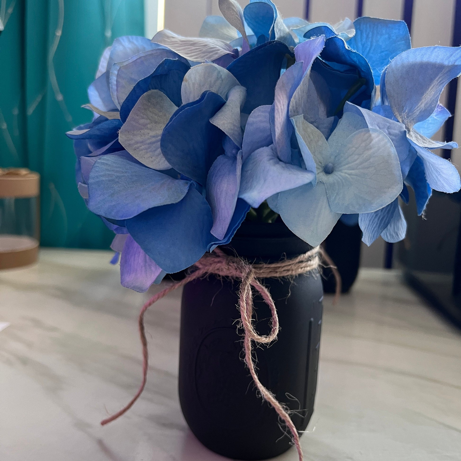 Hand-painted black mason jar with faux blue hydrangea flowers tied with rustic twine. This farmhouse-inspired vase adds a touch of cozy charm to any space, perfect for home decor or special events.