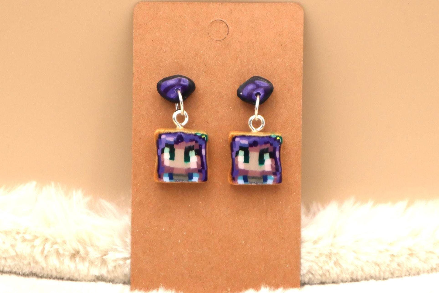 A pair of dangle earrings featuring pixel art of a character's face on a square, toast-like shape. The earrings have purple and light brown accents and silver ear wires.  The pixel art style is reminiscent of video game characters. These handmade polymer clay earrings are a unique and quirky accessory.