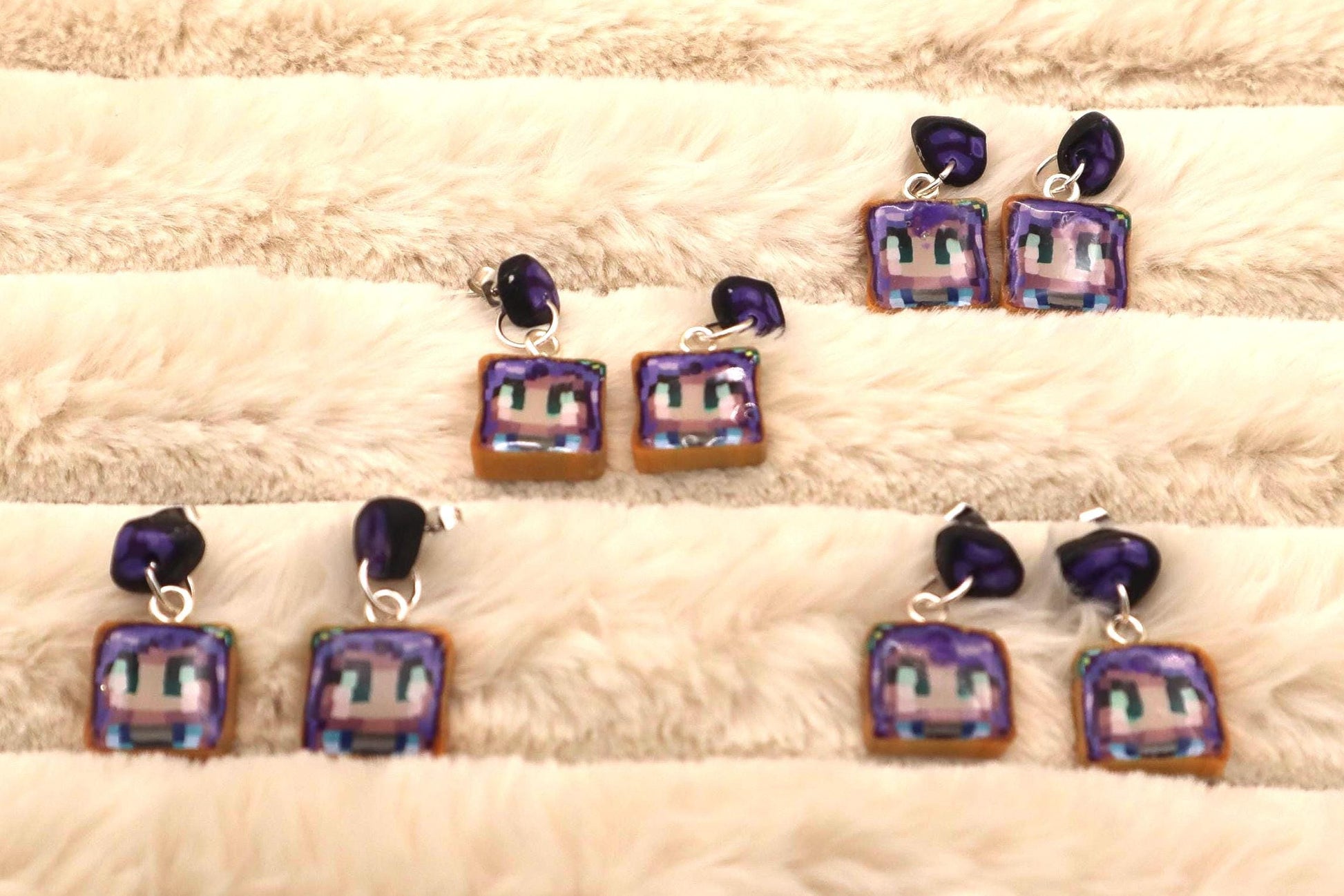 Four pairs of dangle earrings feature pixel art of a character's face on a square, toast-shaped charm.  The charms are brown with purple and white pixel details. The earrings have dark purple beads at the top of the silver ear wires. The earrings are displayed against a light beige fluffy background.