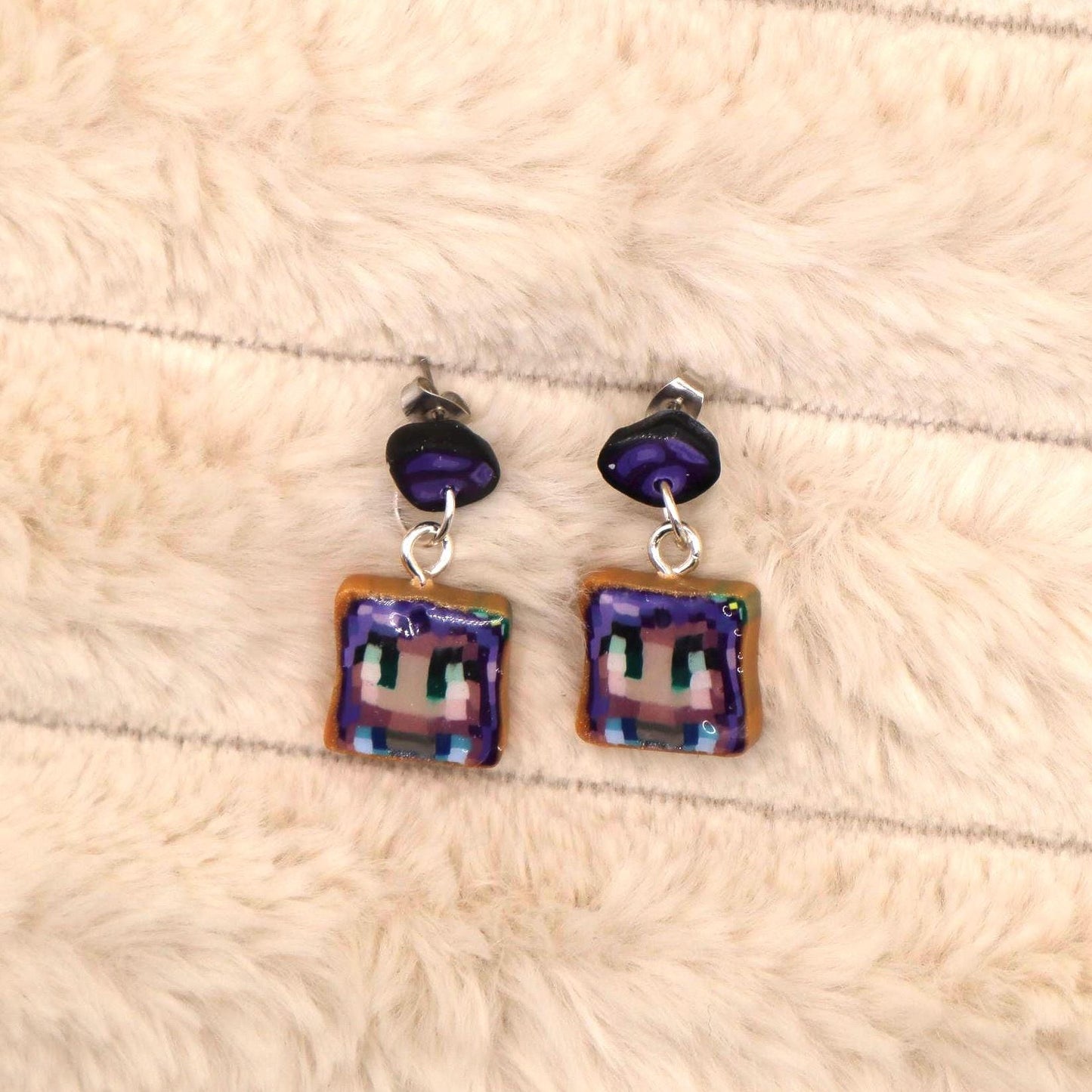 A pair of handmade dangle earrings featuring a pixelated character design on a square, toast-colored base. The character's face is purple with green eyes, and the earrings have dark purple accents.  The earrings are suitable for fans of video games and pixel art. These unique earrings are perfect for adding a touch of playful style to any outfit.