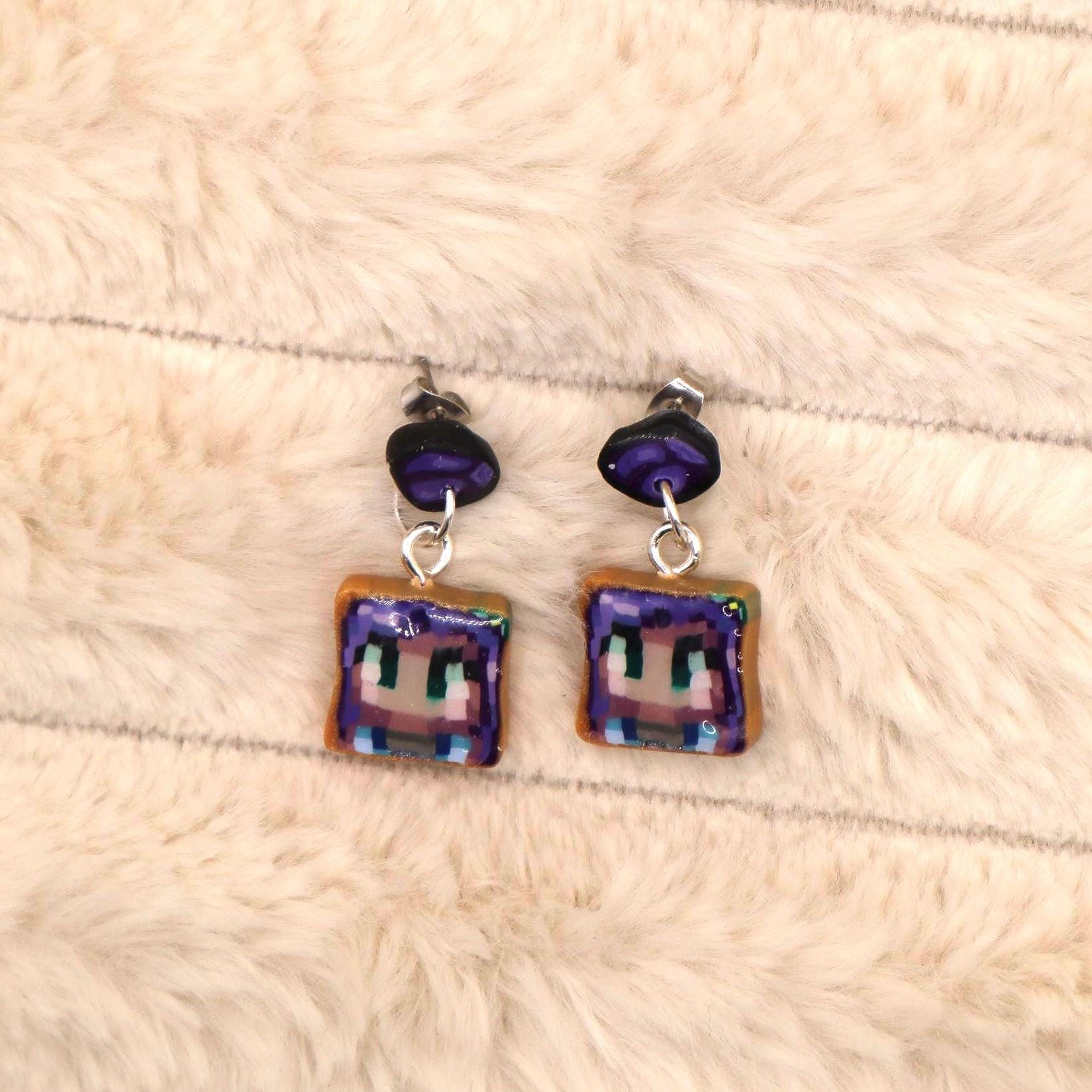 A pair of handmade dangle earrings featuring a pixelated character design on a square, toast-colored base. The character's face is purple with green eyes, and the earrings have dark purple accents.  The earrings are suitable for fans of video games and pixel art. These unique earrings are perfect for adding a touch of playful style to any outfit.