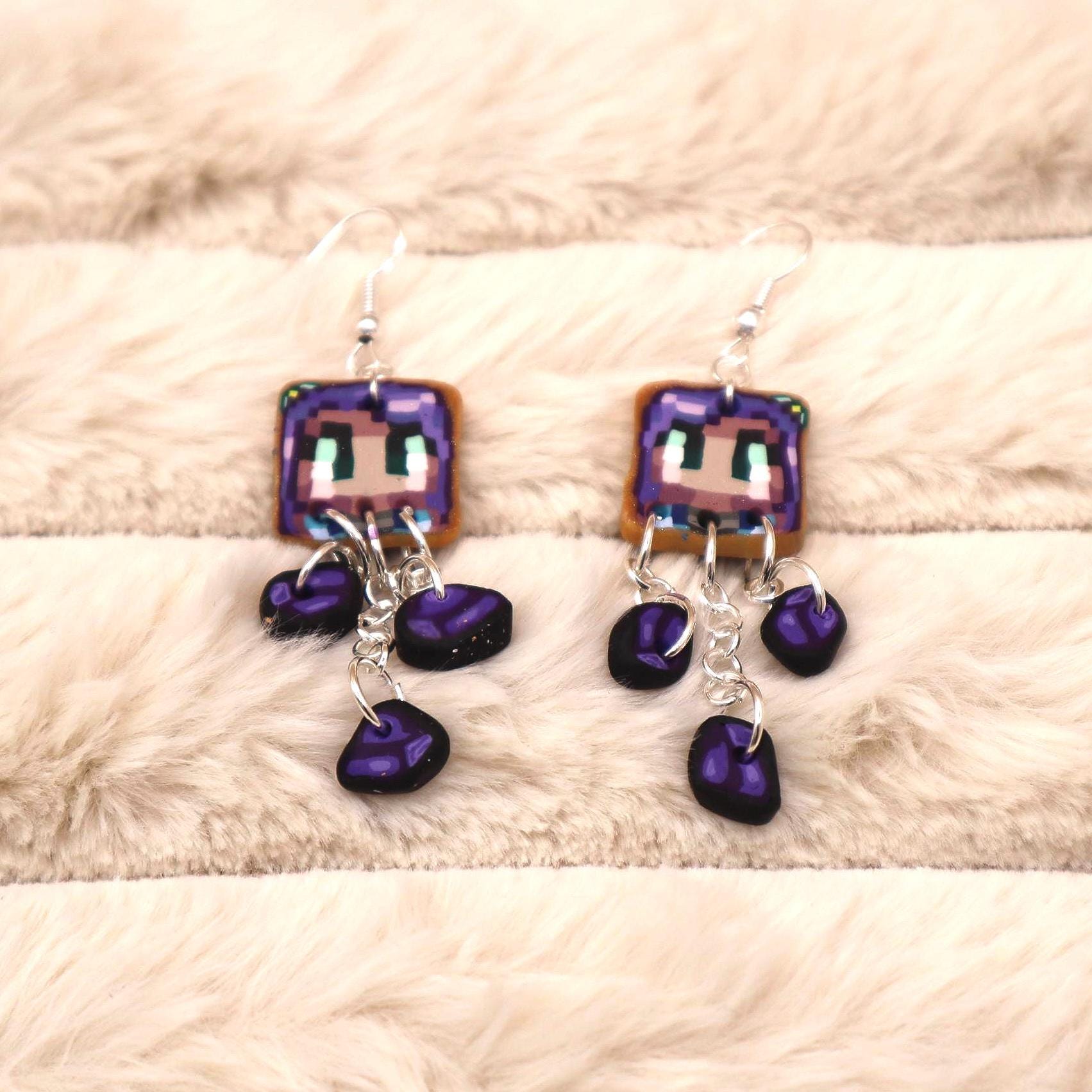 A pair of dangle earrings feature a pixelated character image with purple hair and a light brown complexion.  The square-shaped image is suspended from silver chains, with three dark purple teardrop-shaped beads hanging below. The earrings have a Minecraft-inspired design aesthetic.