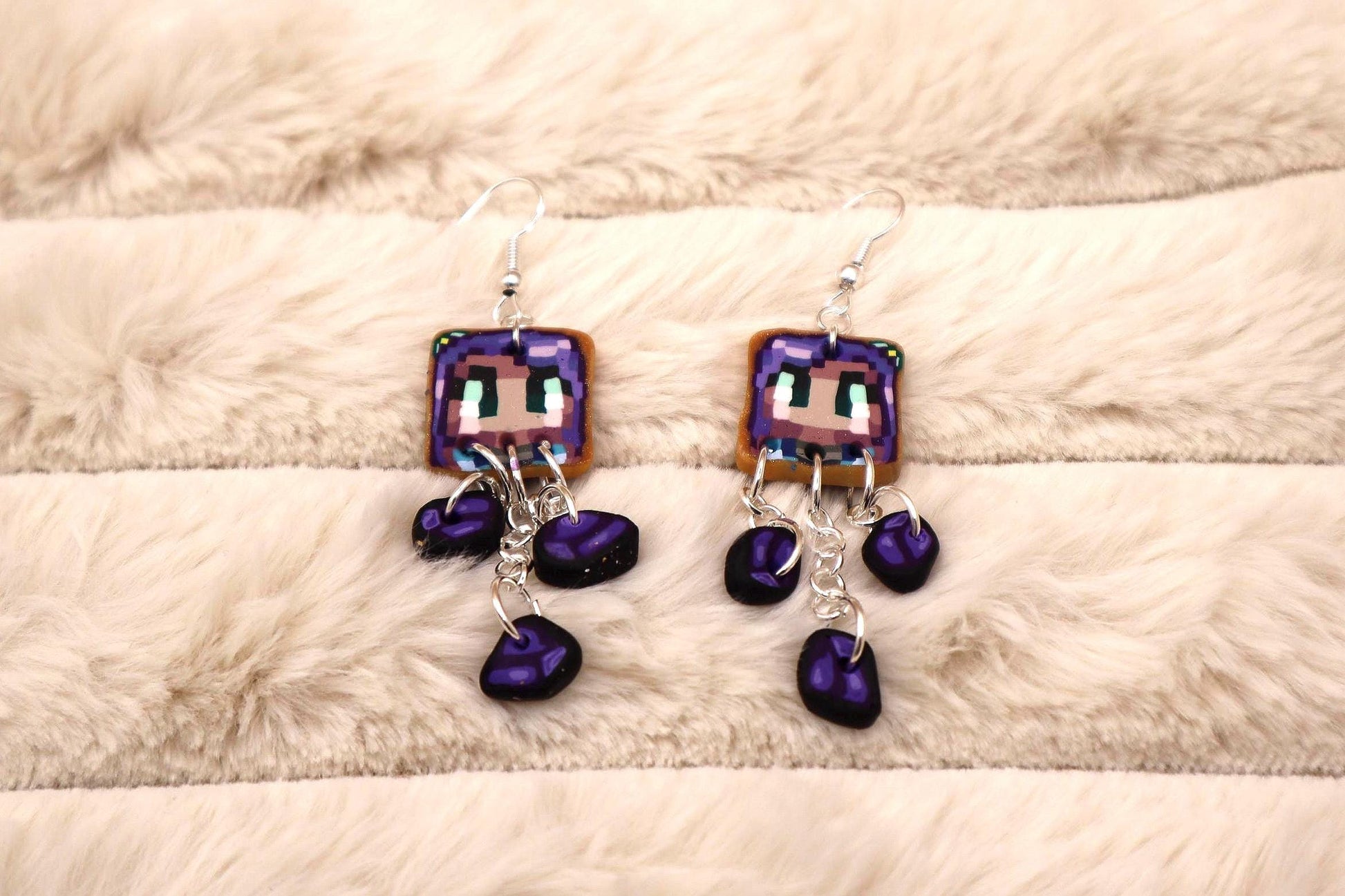 Pixel art earrings featuring a Minecraft-style character face on a square-shaped charm.  The charms are light brown with purple and light blue details. Three dark purple beads dangle from each earring, attached by silver chains. The earrings have silver hooks.