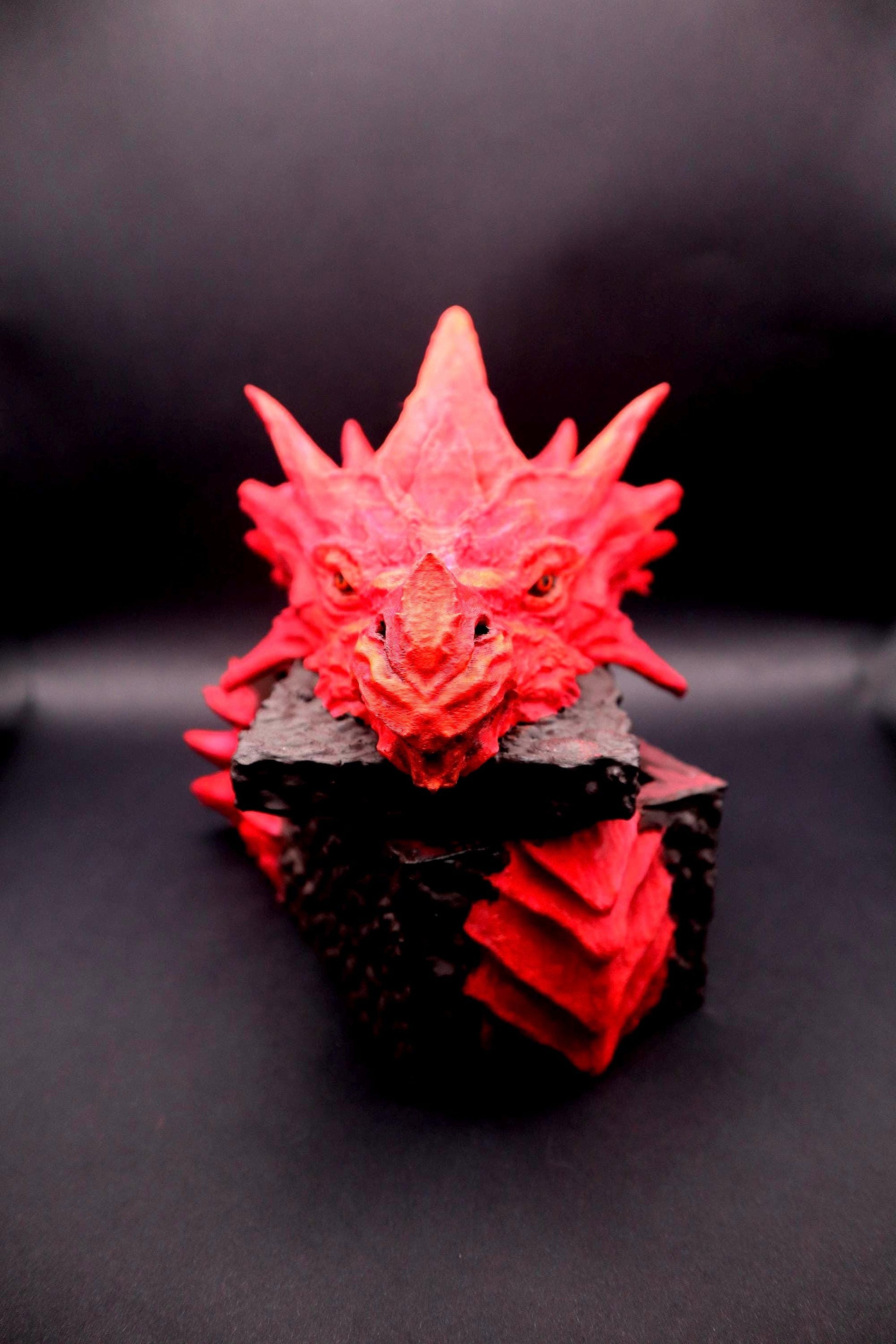 A red dragon head sculpture with black textured base. The dragon has a fierce expression with sharp teeth and claws. Red dragon-themed dice case with resin dice set, perfect for DnD and RPG players. Stylish fantasy dice holder and storage box for tabletop gaming sessions.
