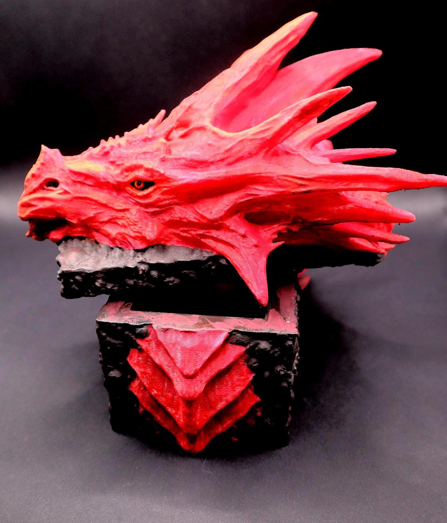 A red dragon head sculpture with a black and red box. The dragon head is open and the box is closed. The dragon head is detailed with scales and teeth. The box has a black base with red accents. Red dragon-themed dice case with resin dice set, perfect for DnD and RPG players. Stylish fantasy dice holder and storage box for tabletop gaming sessions.