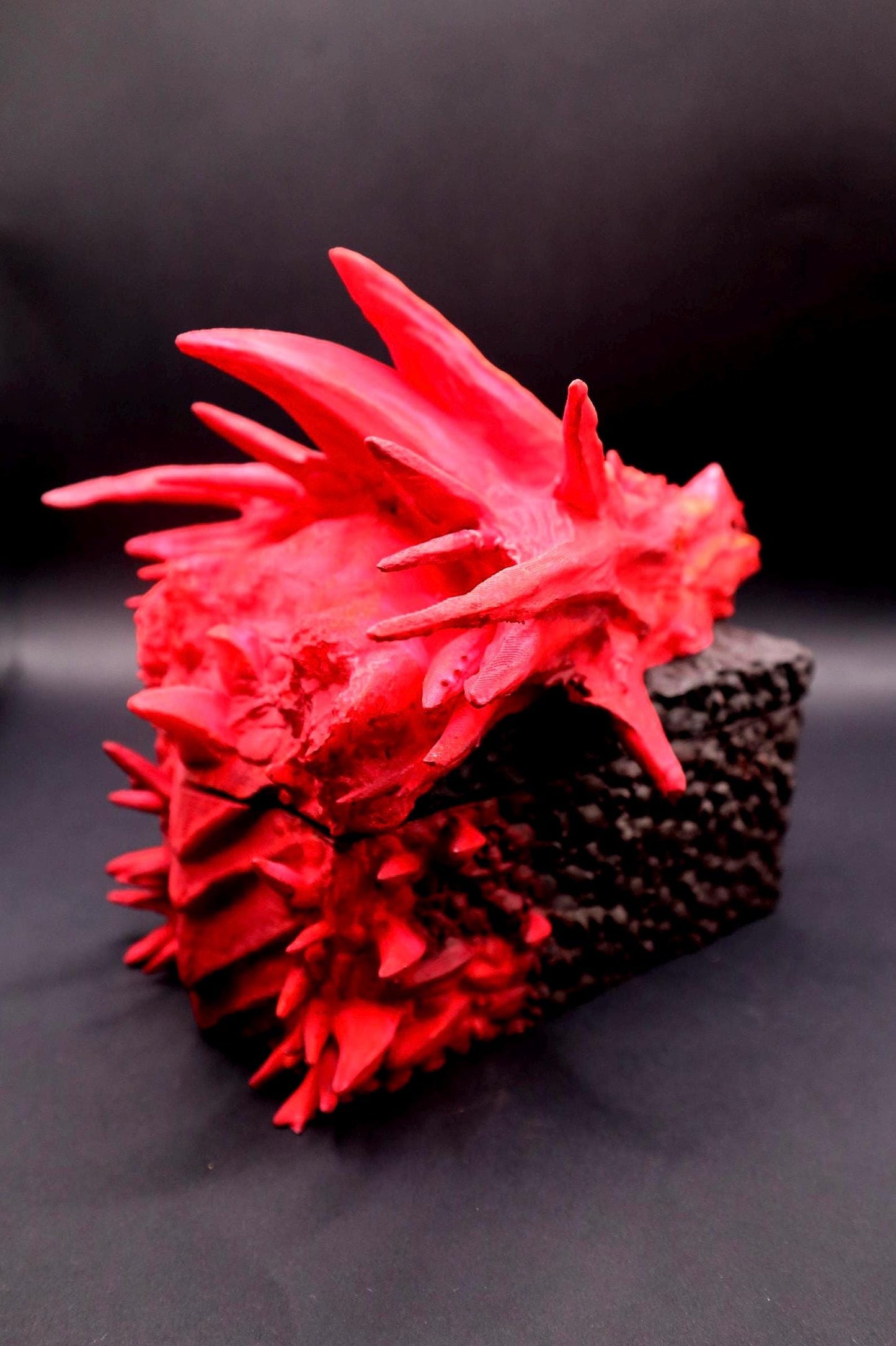 A red dragon sculpture with a textured surface. The dragon is positioned on a black, textured base. Red dragon-themed dice case with resin dice set, perfect for DnD and RPG players. Stylish fantasy dice holder and storage box for tabletop gaming sessions.