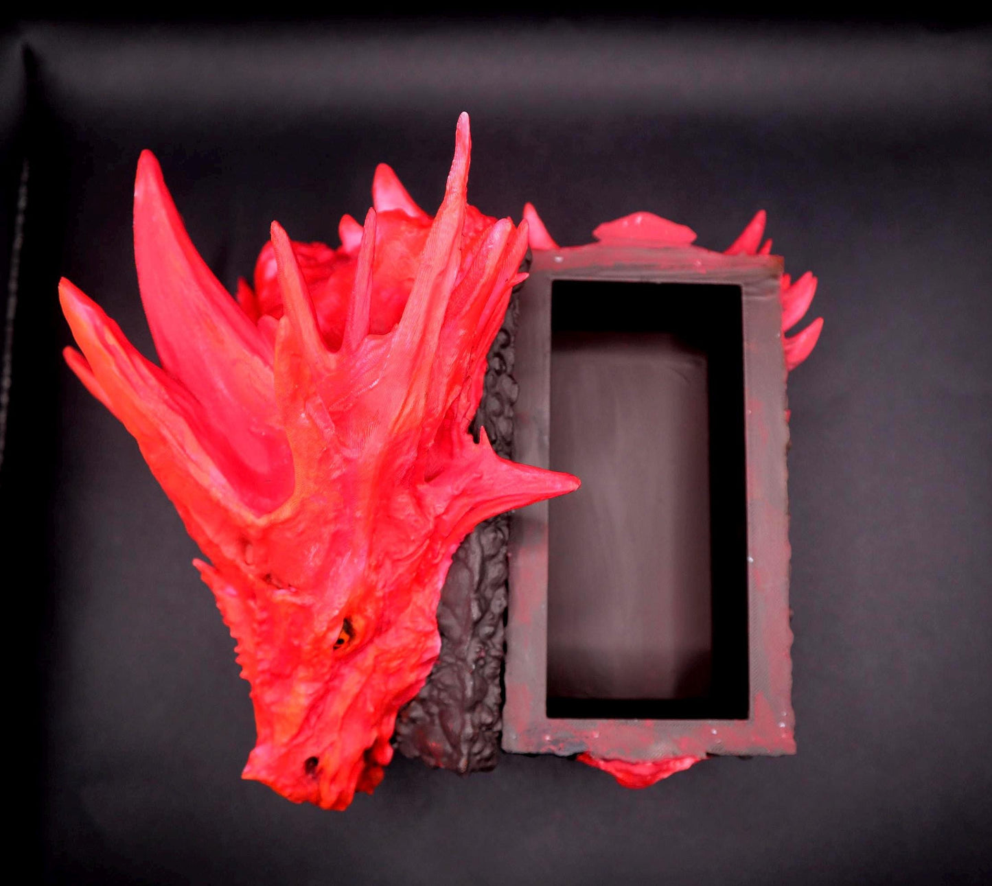 A red dragon head sculpture with an open box behind it. The dragon head is made of a textured material and has a detailed design. The box is made of a dark brown material and has a simple design. Red dragon-themed dice case with resin dice set, perfect for DnD and RPG players. Stylish fantasy dice holder and storage box for tabletop gaming sessions.