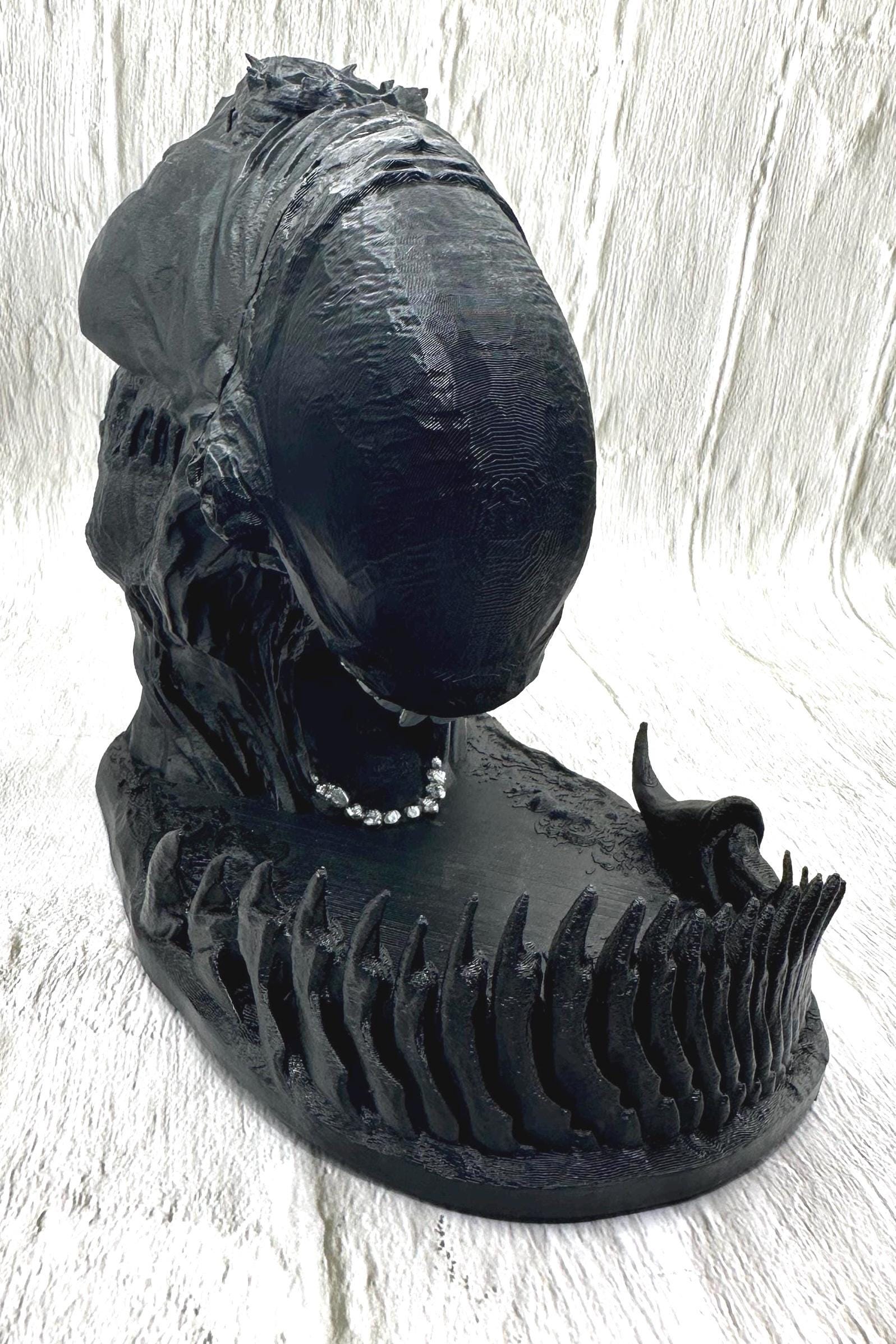 A black 3D printed alien dice tower with silver teeth. The tower is sitting on a black base with a tail around the edge to catch the dice dropped in the slot at the top of his head.