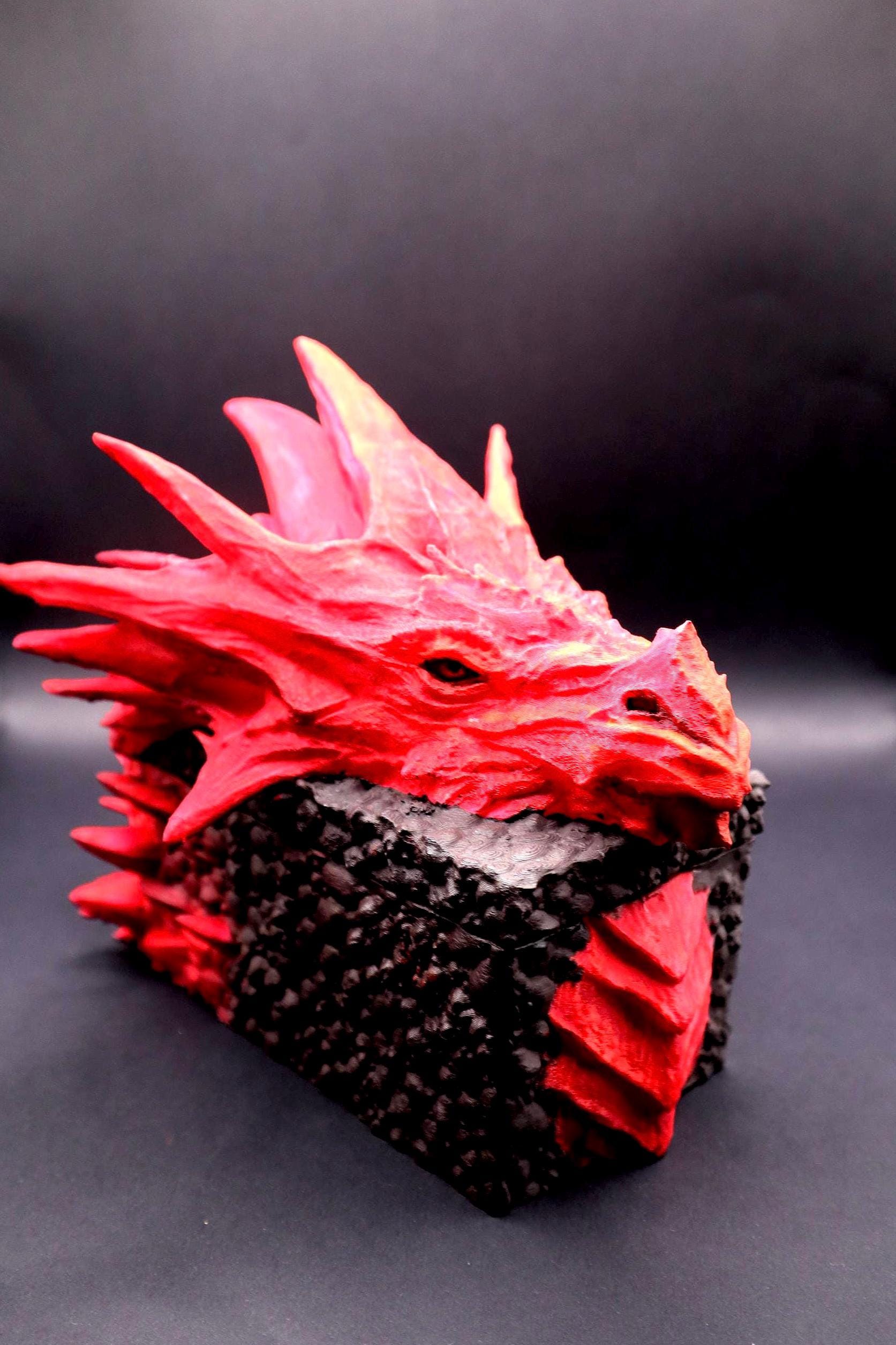 A red dragon head sculpture with a black textured base. The dragon has a detailed face with a yellow eye and a textured, scaly body. Red dragon-themed dice case with resin dice set, perfect for DnD and RPG players. Stylish fantasy dice holder and storage box for tabletop gaming sessions.