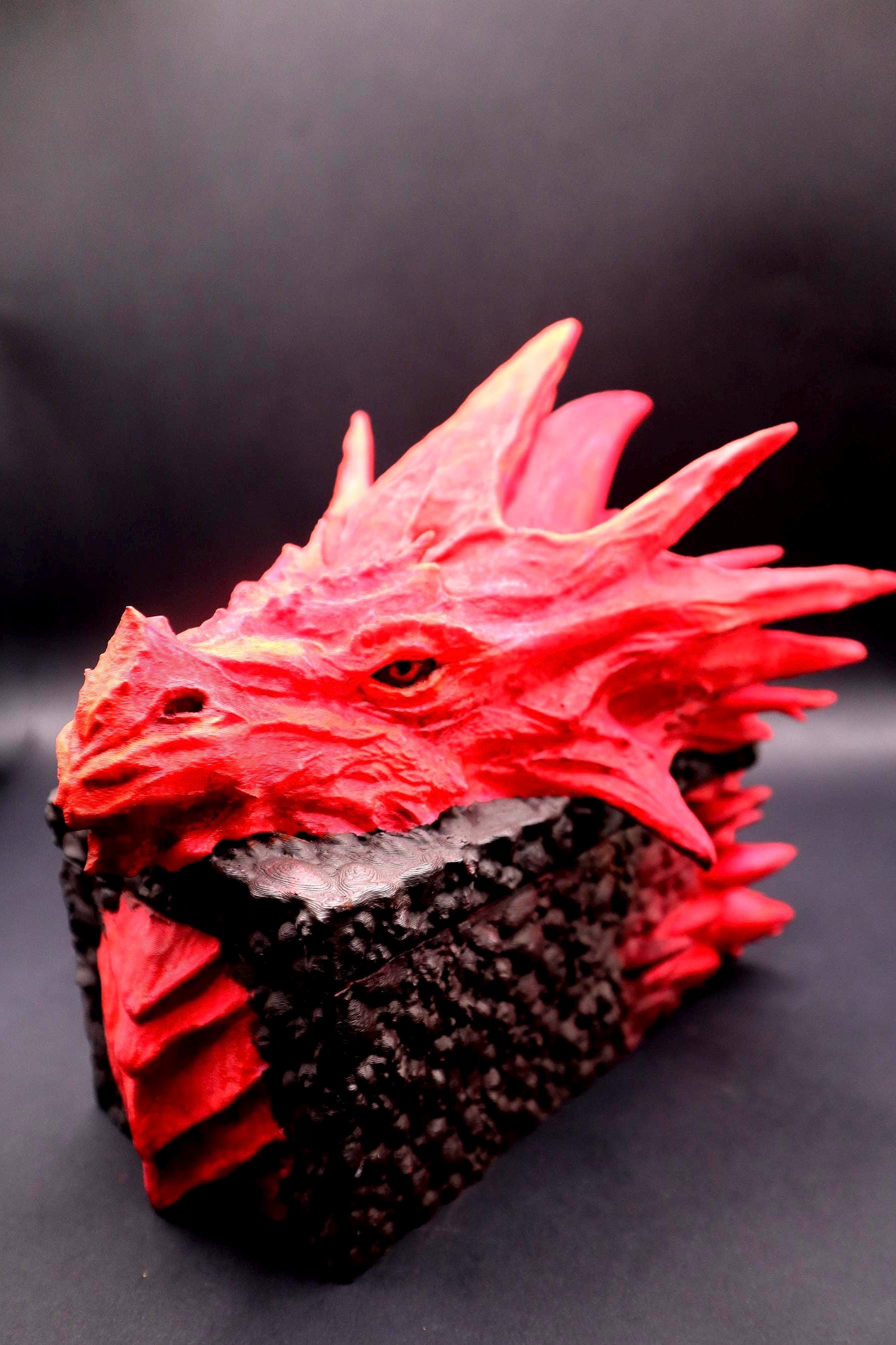 A red dragon head sculpture with a textured surface and a black base. The dragon's eyes are painted red and the sculpture is made of a hard material. Red dragon-themed dice case with resin dice set, perfect for DnD and RPG players. Stylish fantasy dice holder and storage box for tabletop gaming sessions.