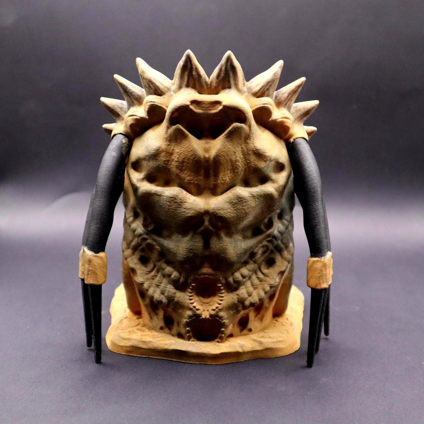 A Predator dice tower with a tan and brown color scheme. The tower features a detailed, textured design with a wide open mouth revealing pink gums and white teeth. The tower has black dreadlocks and a spiky head crest, showcasing the iconic alien creature inspired by the Predator movie franchise.
