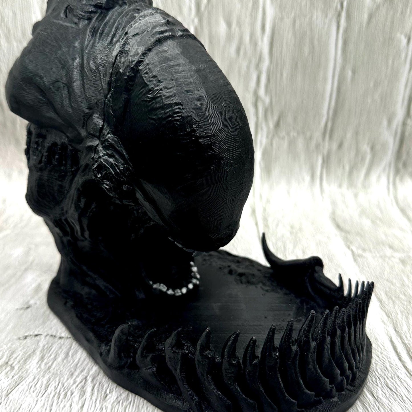 A black 3D printed alien dice tower with silver teeth. The tower is sitting on a black base with a tail around the edge to catch the dice dropped in the slot at the top of his head.
