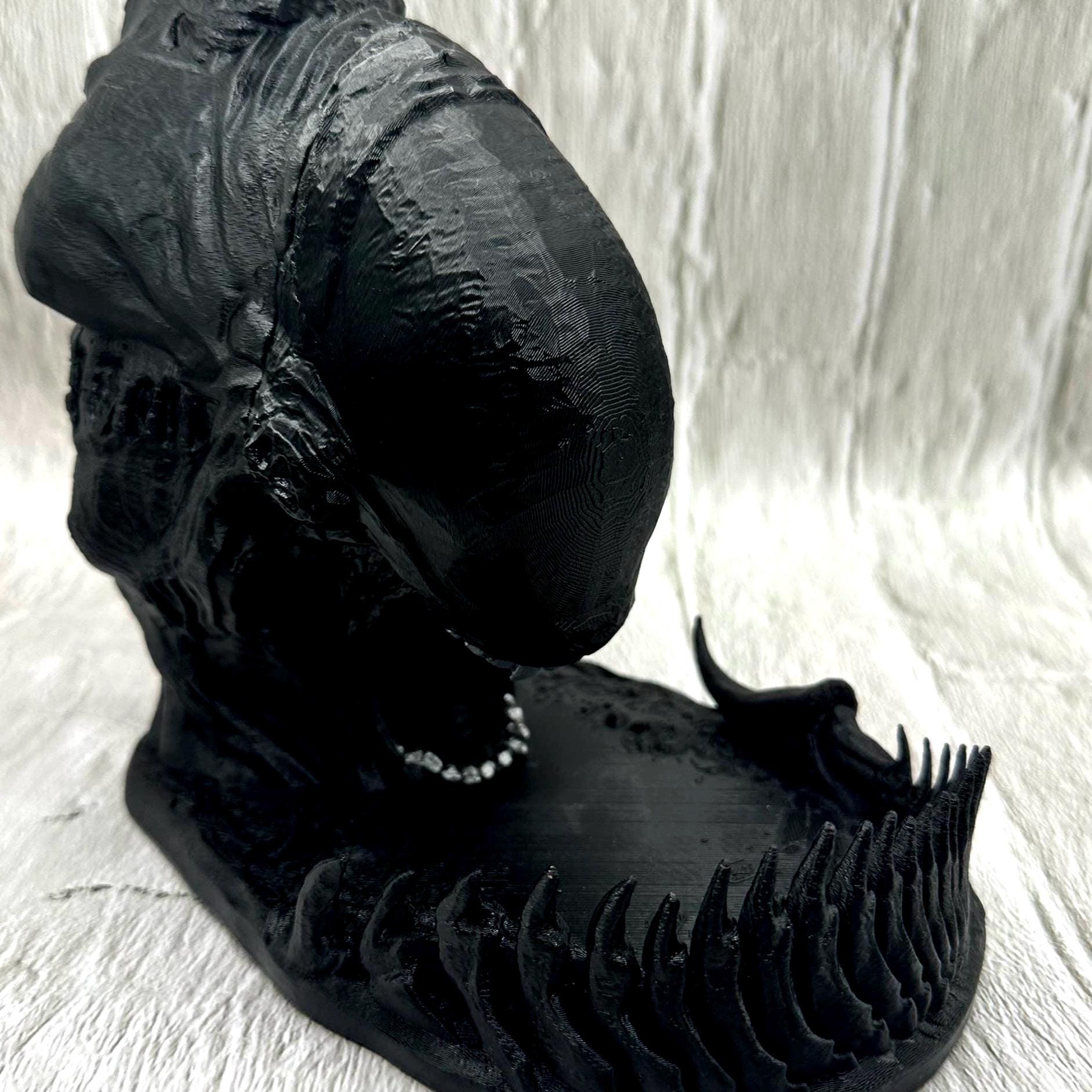 A black 3D printed alien dice tower with silver teeth. The tower is sitting on a black base with a tail around the edge to catch the dice dropped in the slot at the top of his head.